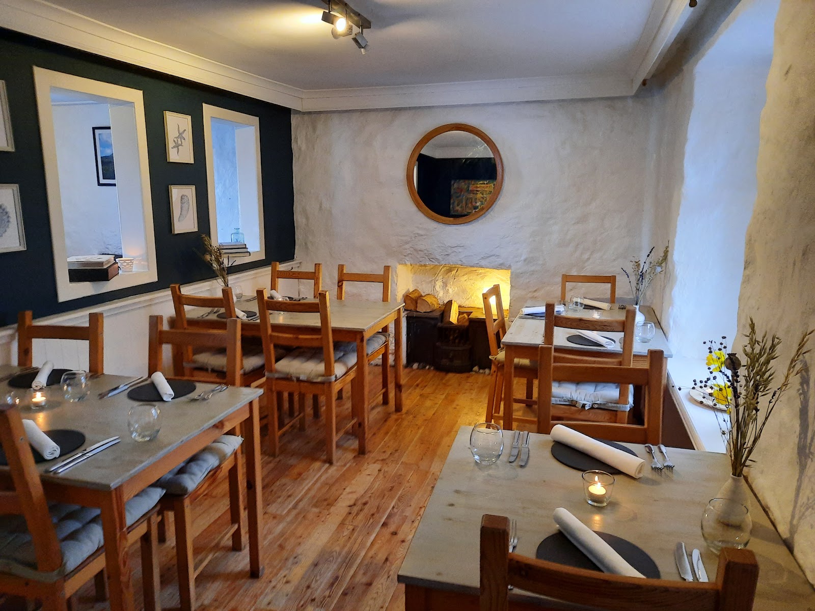 The dining room at The Whitehouse Restaurant in Lochaline, wooden tables laid with candles, white stone walls, a round mirror and a log fireplace.
