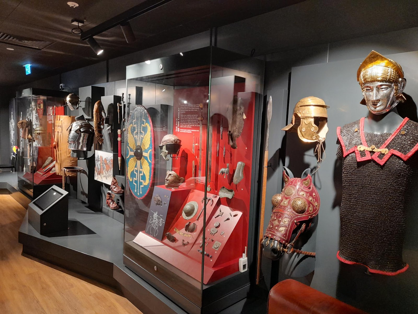 Inside the Trimontium Museum: Roman cavalry armour, helmets, a painted shield and a silver face-mask helmet in dark cases lit red.
