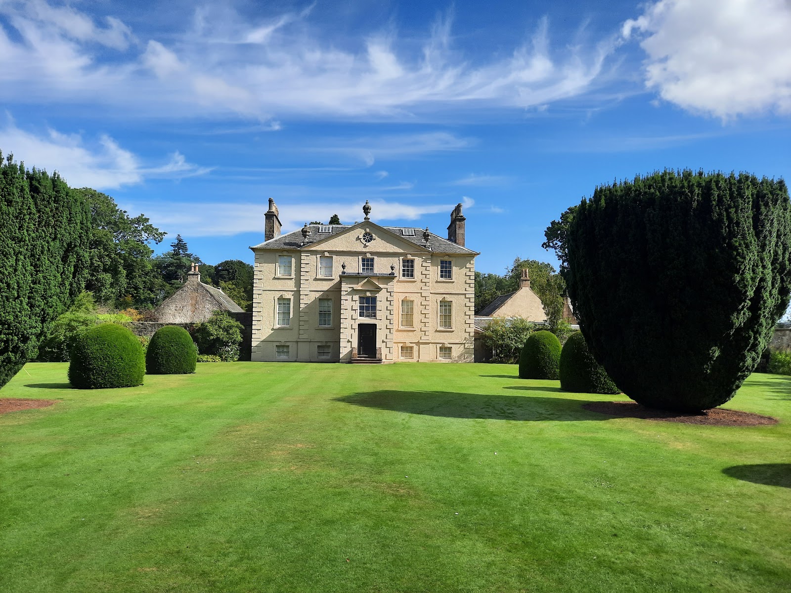 Greenbank House, a pale Georgian mansion, across a wide lawn framed by clipped yews under a blue sky.