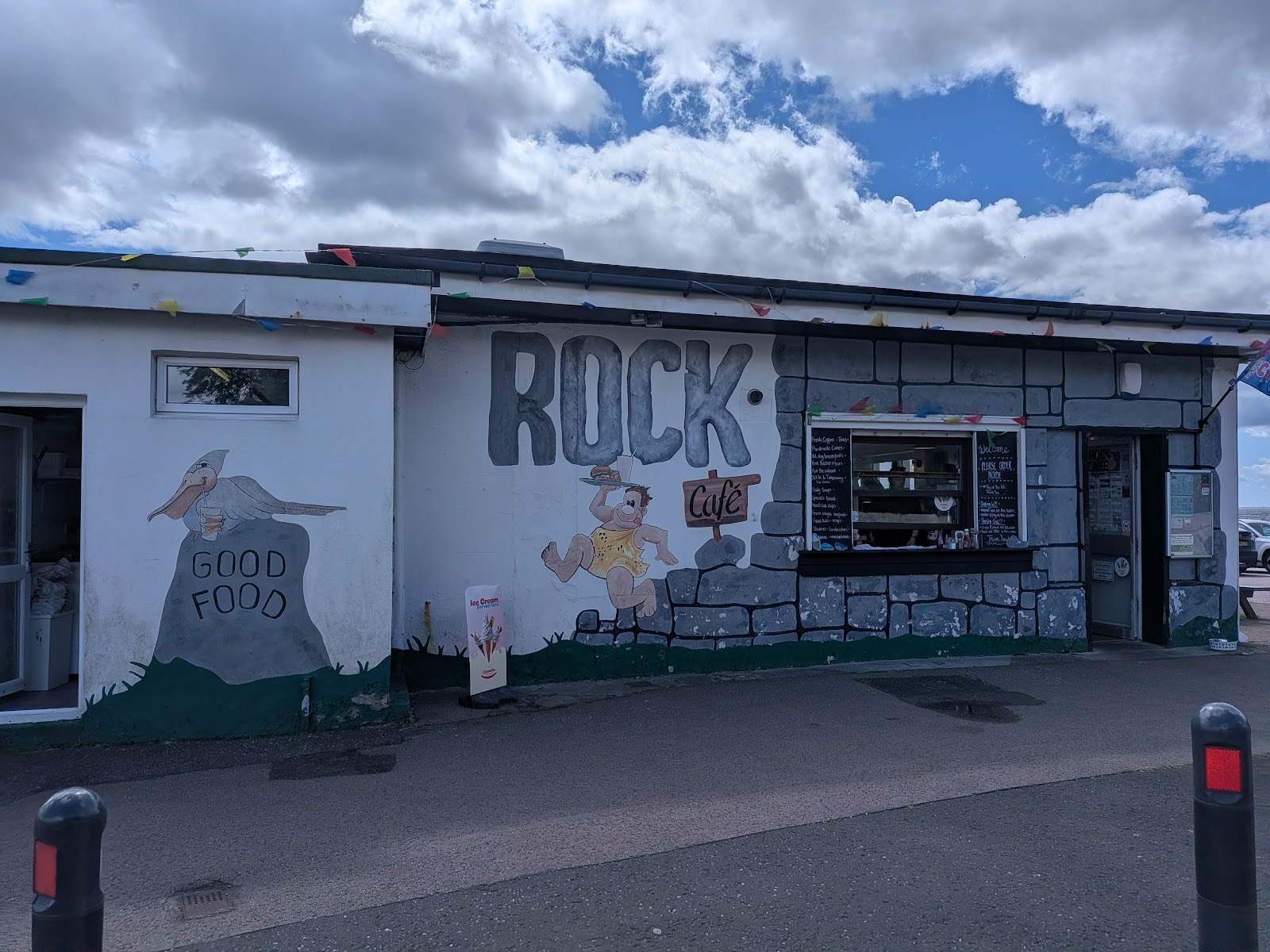 The front of Rock Cafe on West Bay in Dunoon, painted with a big ROCK sign, a caveman carrying a tray and a stone wall, bunting along the roof.