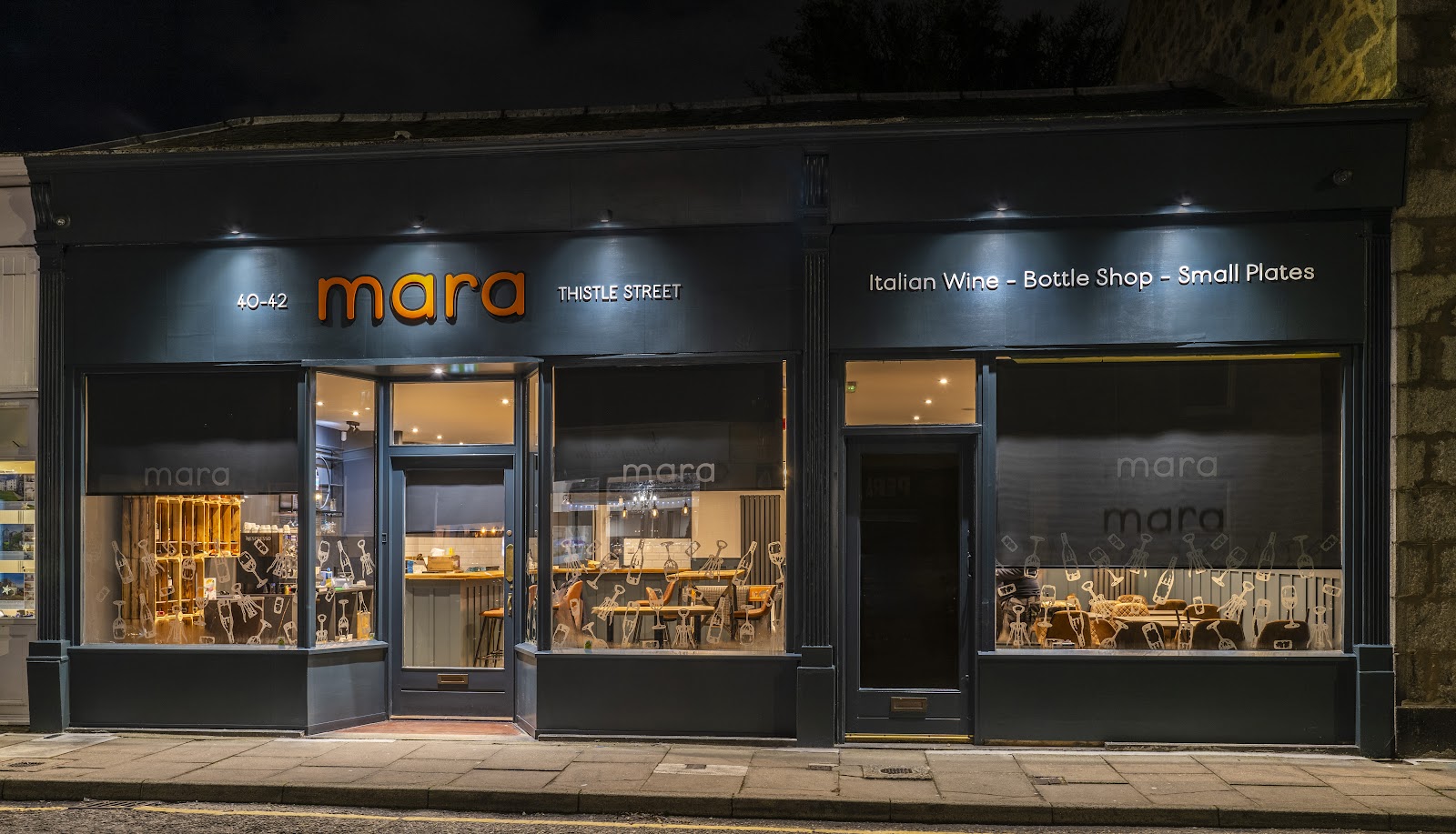The front of Mara on Thistle Street at night, its dark shopfront lit and signed Italian Wine, Bottle Shop, Small Plates, with tables and wine shelves seen through the windows.
