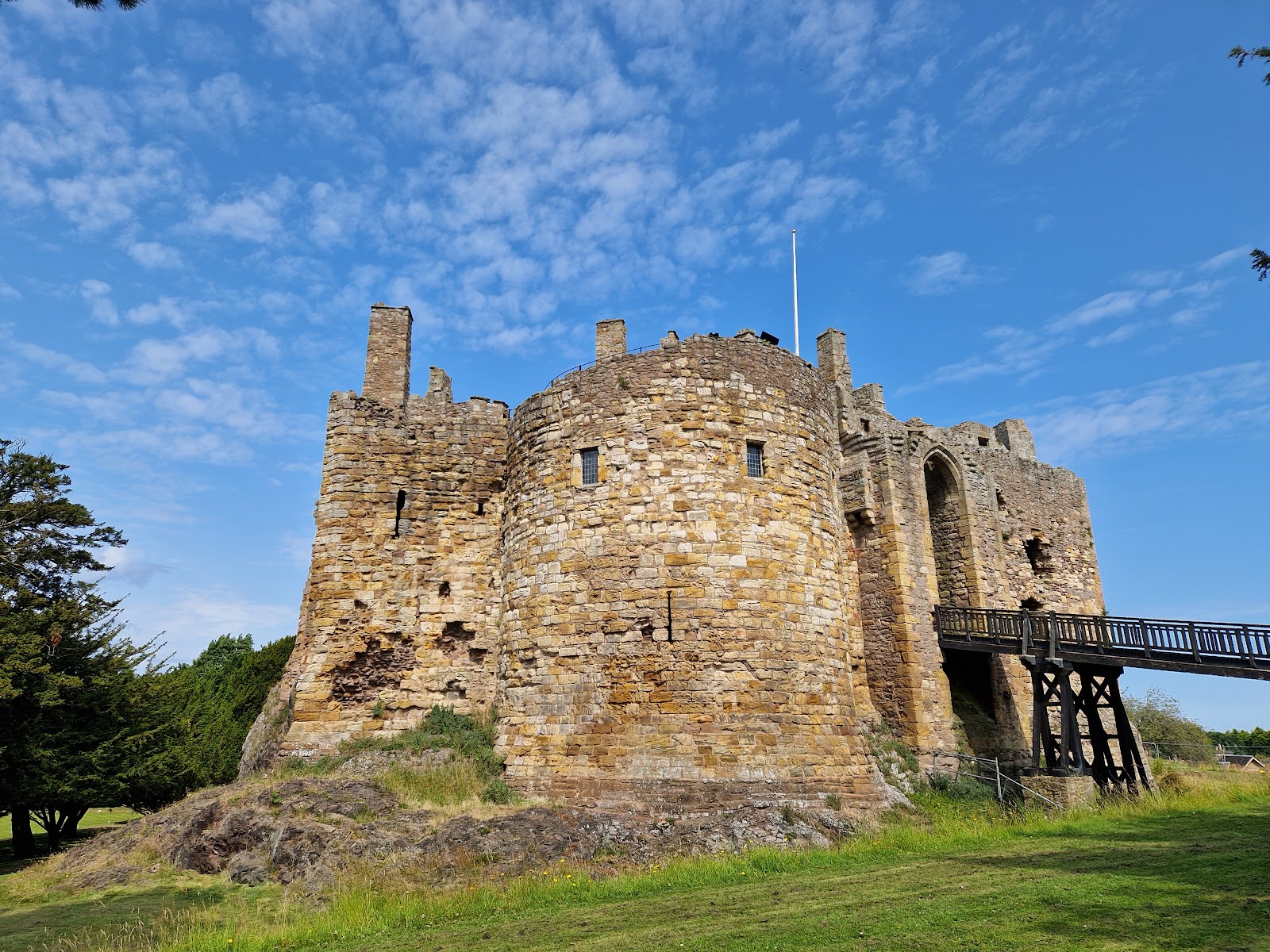 Dirleton Castle, a sandstone ruin on its rock with a wooden bridge to the gate under a blue sky.