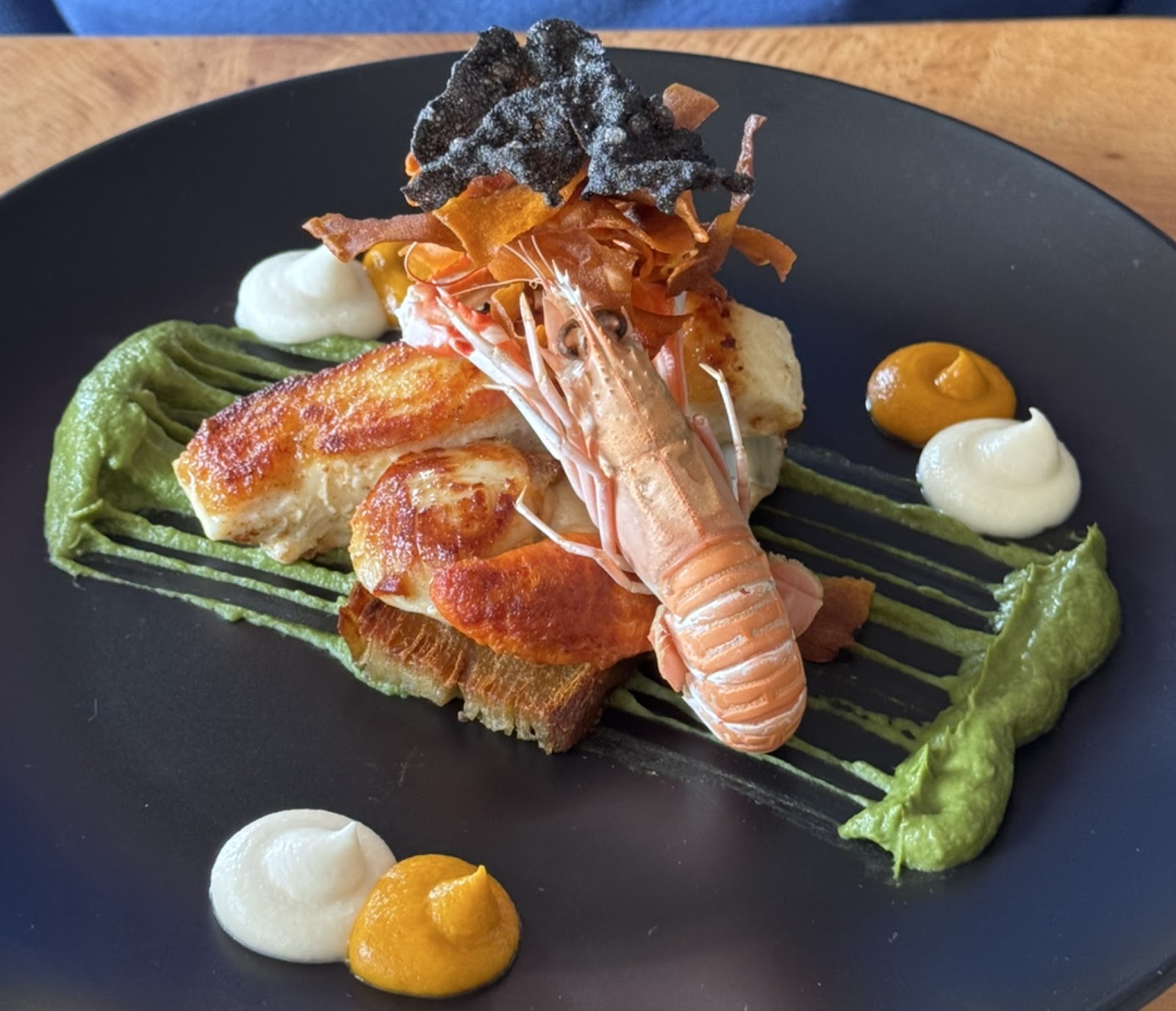 A plate at North Harbour Bistro: a langoustine laid over fish on toast, with sauces and crisp seaweed on a dark plate.