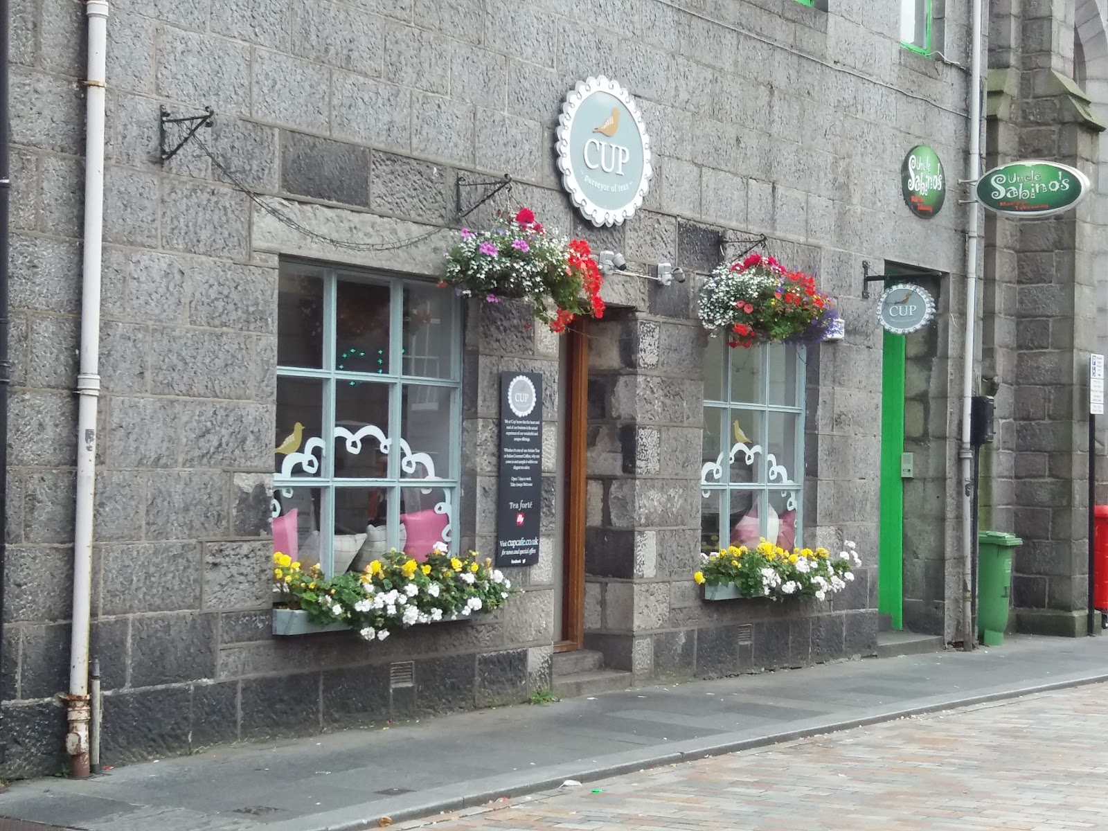 The granite front of Cup on Little Belmont Street, with hanging baskets and window boxes of flowers.