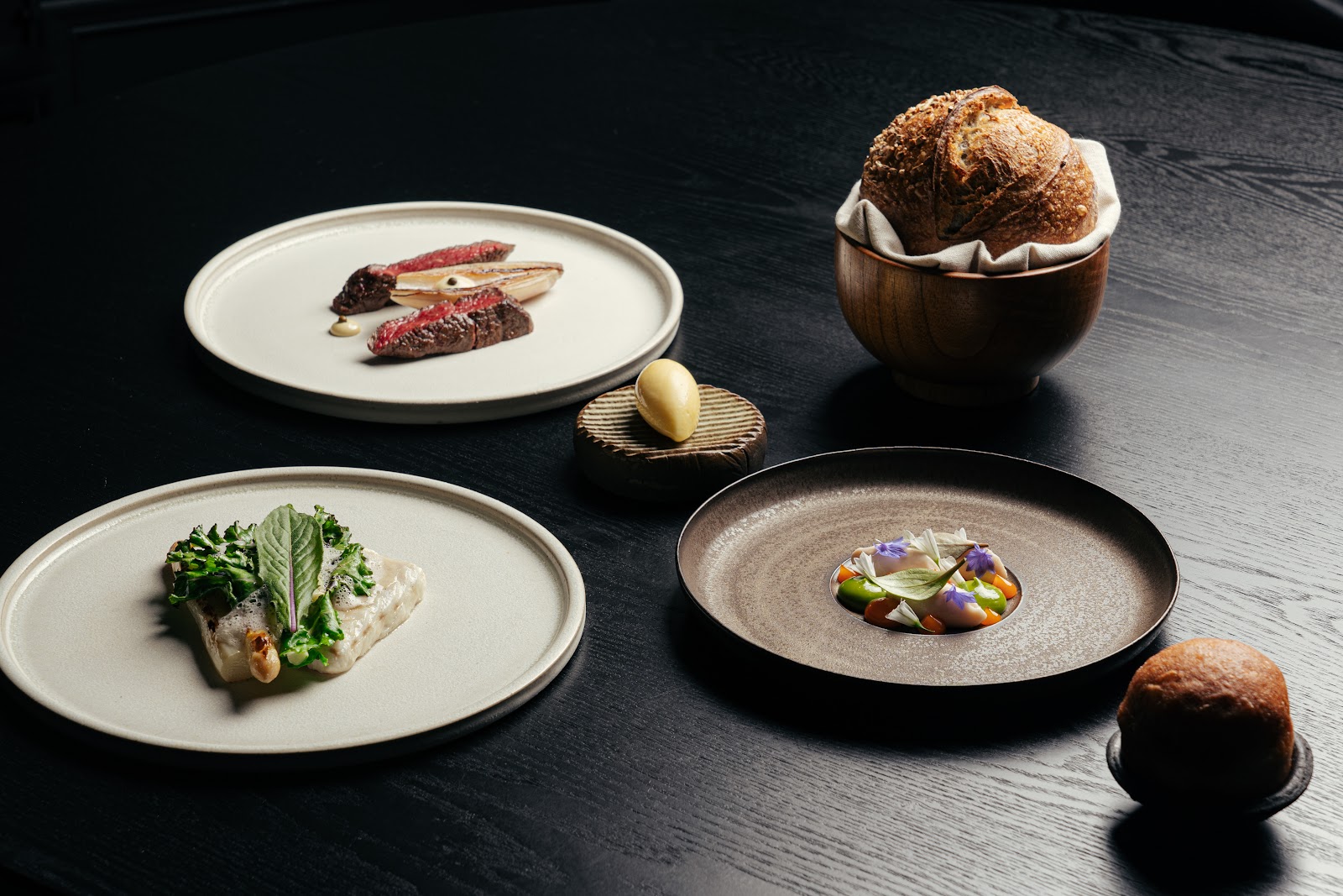 Three courses from Cardinal's tasting menu on a dark table, with a loaf of bread in a wooden bowl and a small dish of butter.