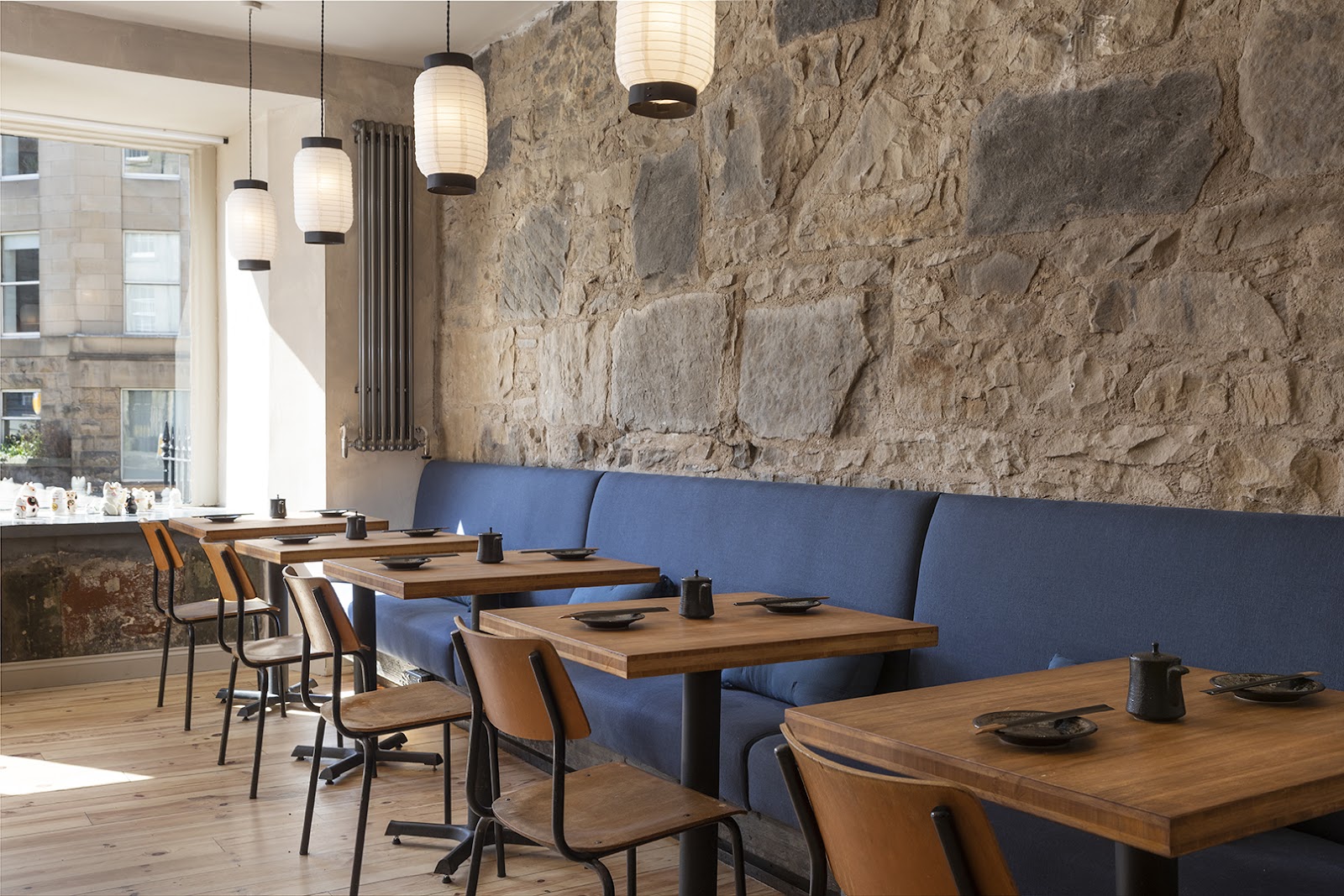 Lucky Yu's dining room, a blue banquette and wooden tables along a rough stone wall under white paper lanterns, with lucky cats in the window.