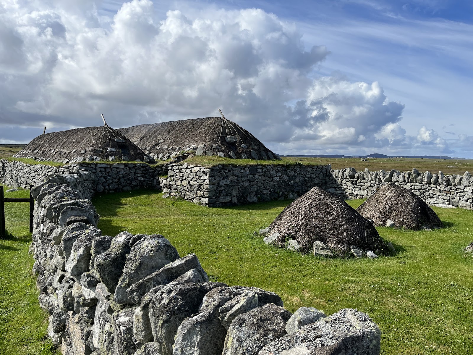 The thatched blackhouses at Arnol behind a drystone wall on green grass, two stacks of thatch in the yard under a big sky of cloud.