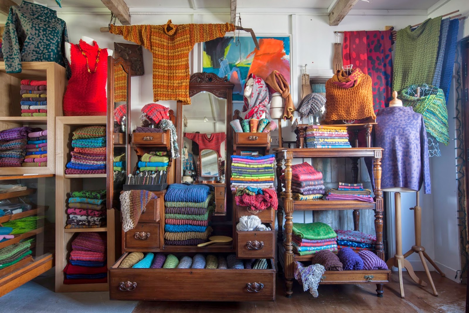 Inside the Nielanell studio shop in Hoswick: colourful knitwear folded on an old wooden dresser and shelves, with jumpers and wraps hung on mannequins and a painting on the wall.