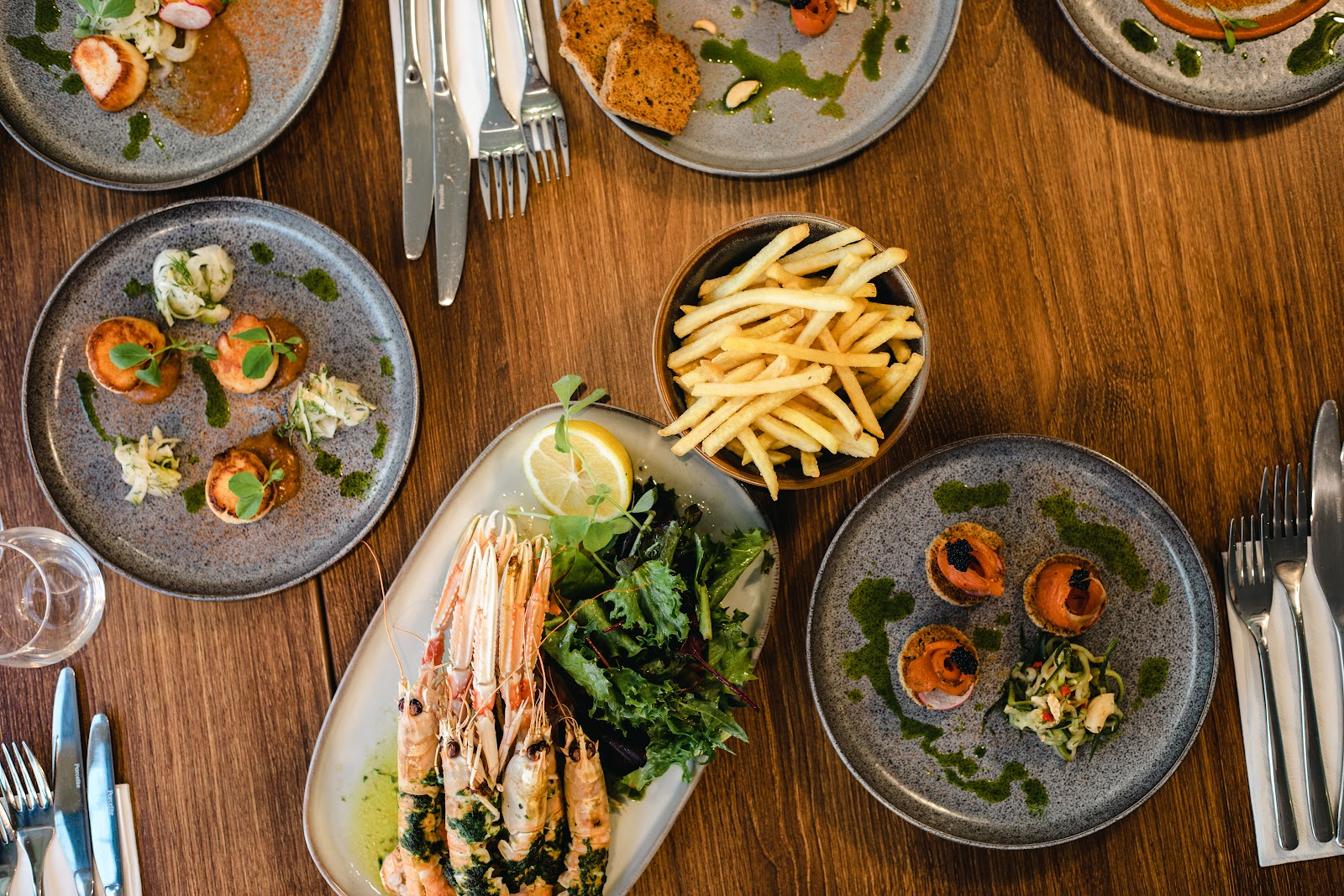 Plates of seafood at Bistro Verde seen from above on a wooden table: langoustines with salad, scallops, smoked salmon bites and a bowl of fries.