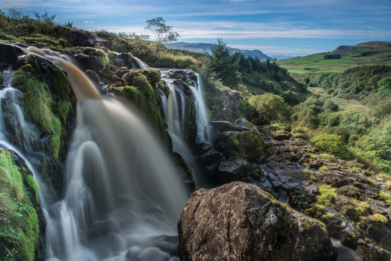 The Endrick Water pouring over mossy rock steps at the top of the Loup of Fintry, with the green valley and hills stretching away beyond.