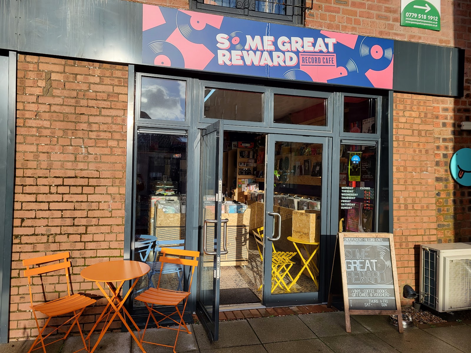 The glass front of Some Great Reward at the Cooperage, its pink and blue sign above the door, with orange chairs and a table outside in the sun.