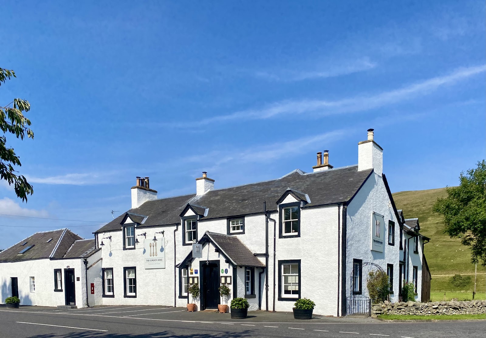 The Gordon Arms in the Yarrow Valley, a long whitewashed coaching inn with dormer windows and tall chimneys under a blue summer sky, green hills rising behind.