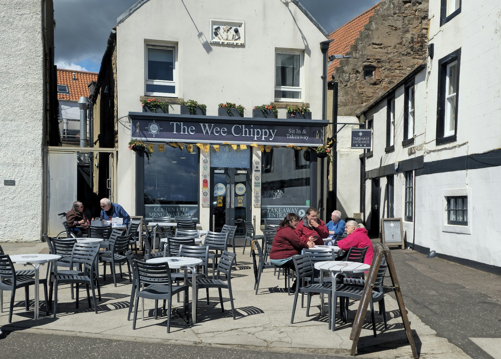 The Wee Chippy on Shore Street, Anstruther, on a sunny day, with people eating at the tables outside its front.