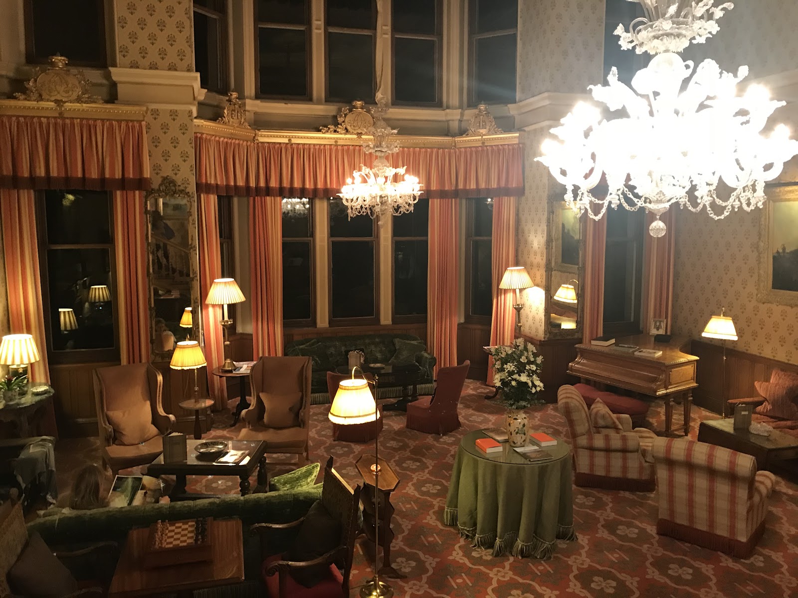 The Great Hall at Inverlochy Castle at night, with glass chandeliers, tall curtained windows, armchairs and a piano.