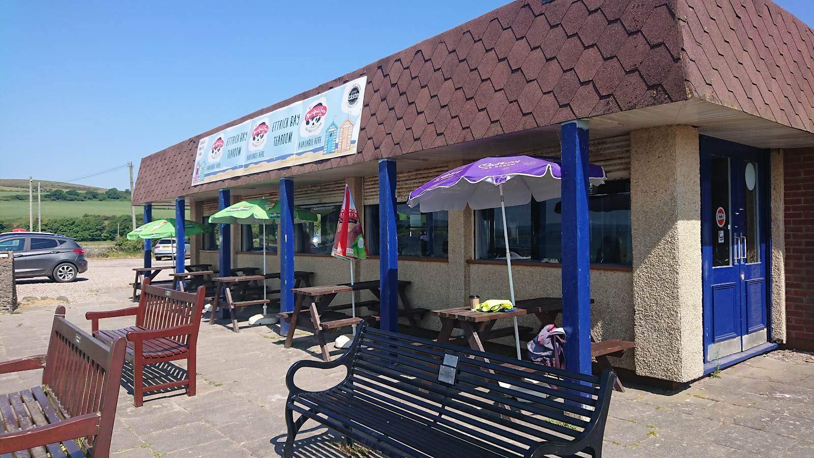 The low brick café at Ettrick Bay on a sunny day, with blue pillars and doors, benches and parasols along the front.