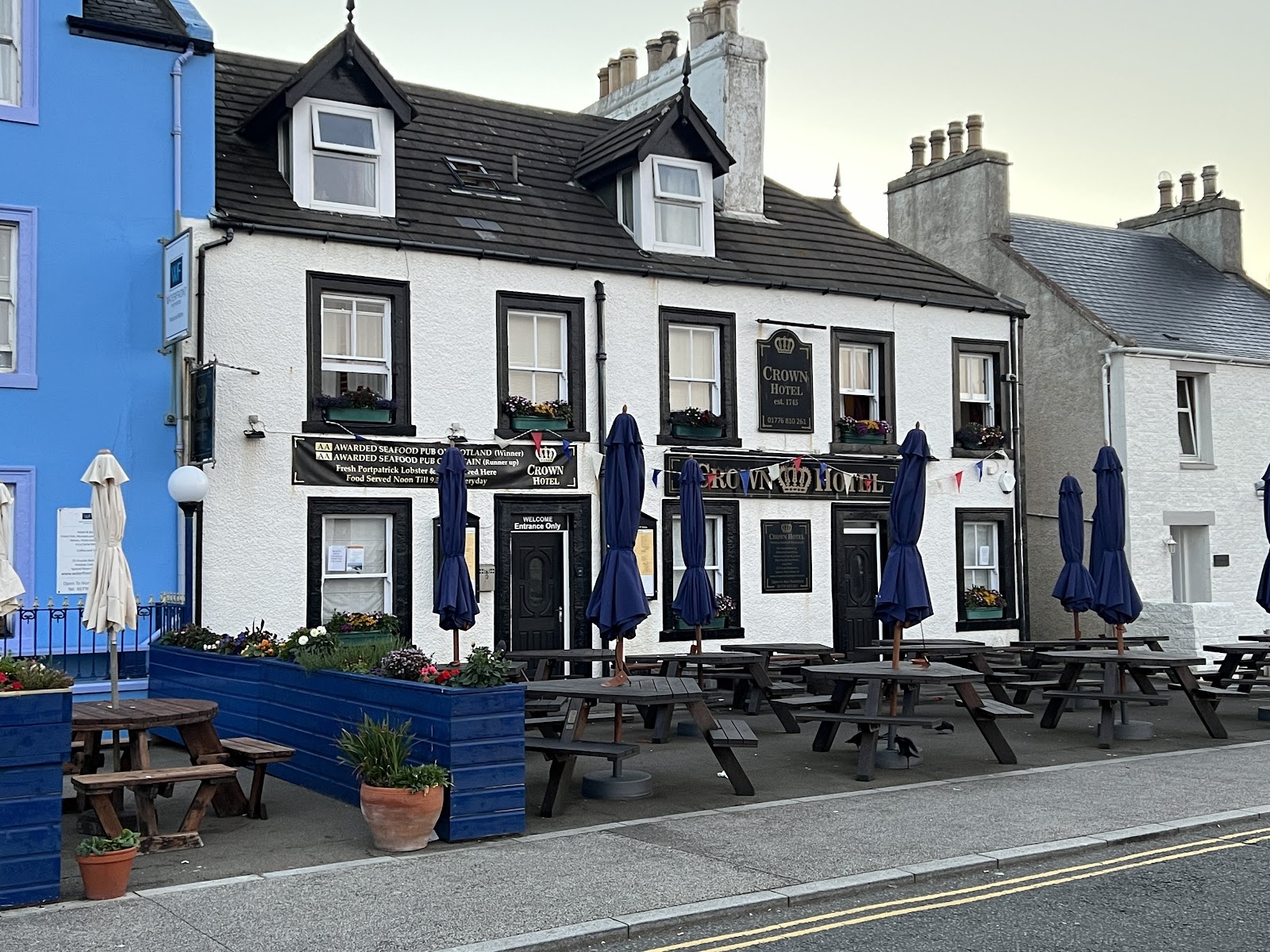 The Crown Hotel's white frontage on North Crescent in Portpatrick, its name over the door, window boxes of flowers and picnic tables out front under furled blue umbrellas.