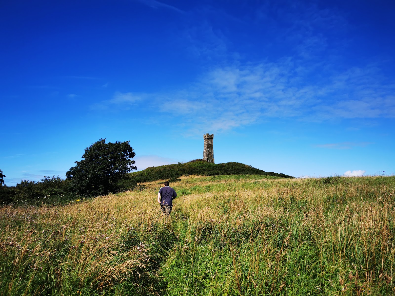 The Agnew Monument, a square castellated stone tower, on the green top of the Tor of Craigoch under a deep blue sky, a walker climbing through the long summer grass towards it.