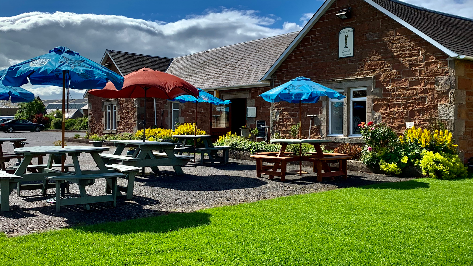 The Caddy Mann, a stone building with a garden of picnic tables under blue parasols on a sunny day.
