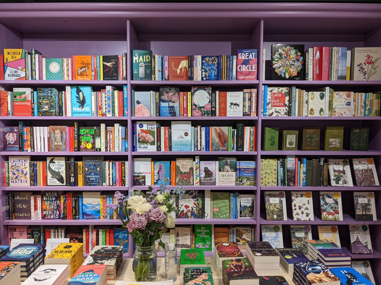 Purple shelves of new books at Night Owl Books in East Linton, with a vase of flowers on the table of books in front.