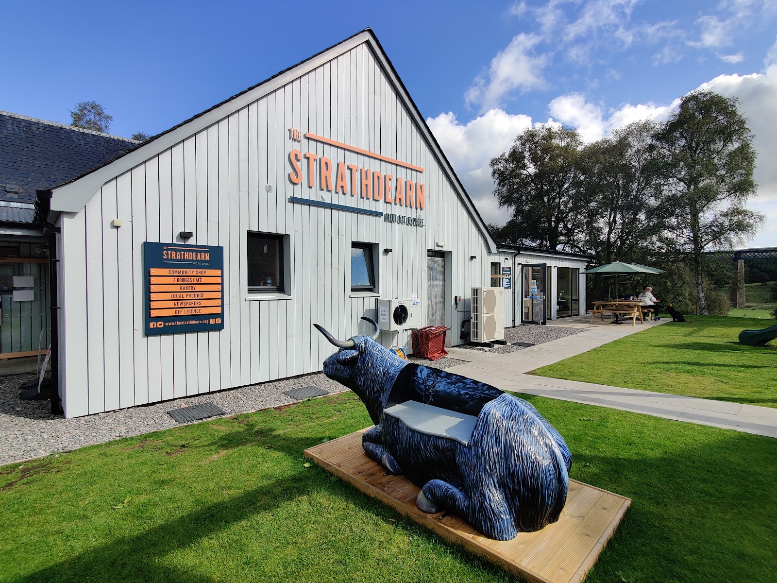 The Strathdearn shop and café in Tomatin, a Highland cow bench on the lawn and the viaduct behind the trees.