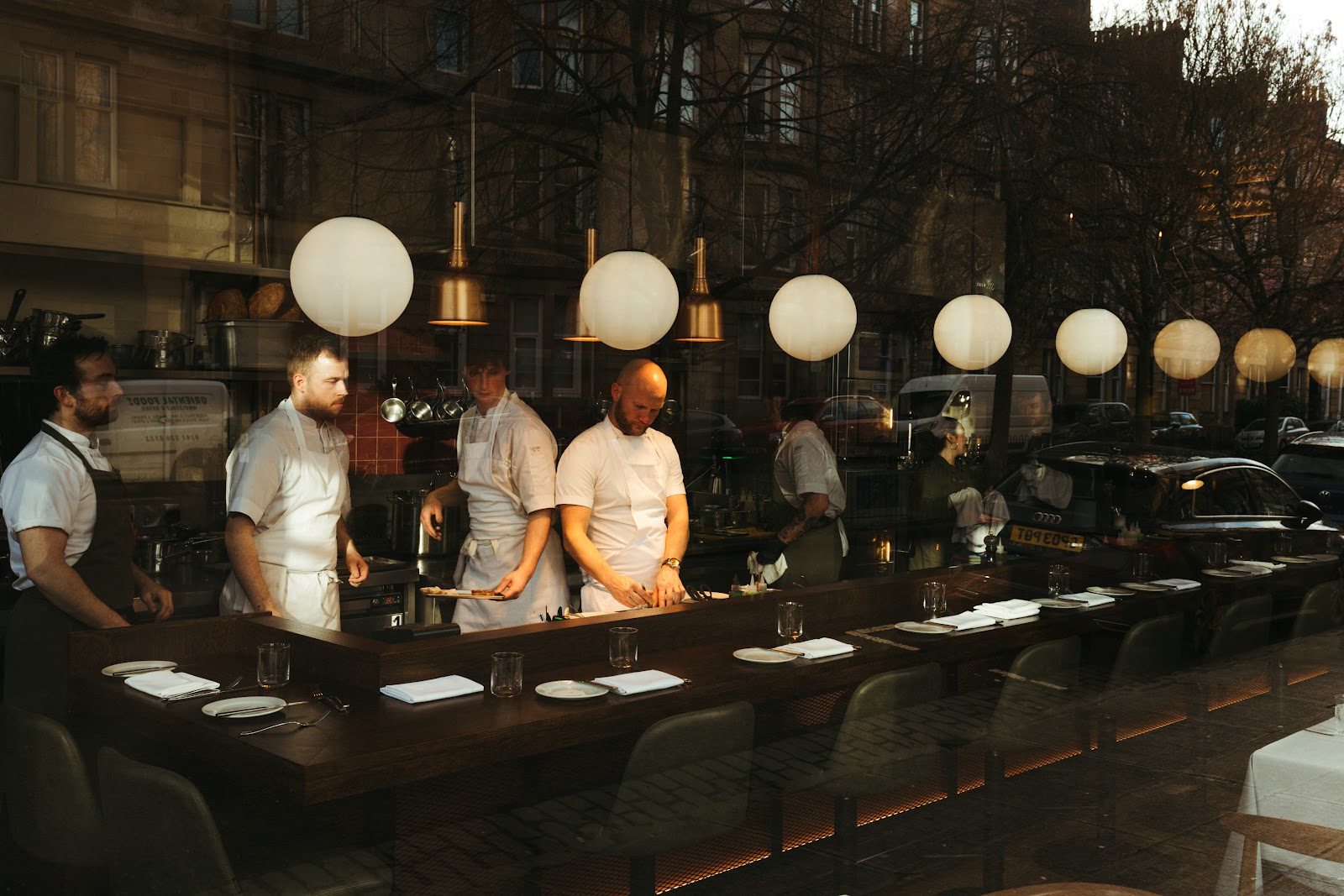 Chefs at work behind the counter of Brett's open kitchen, seen through the window from Great Western Road at dusk.