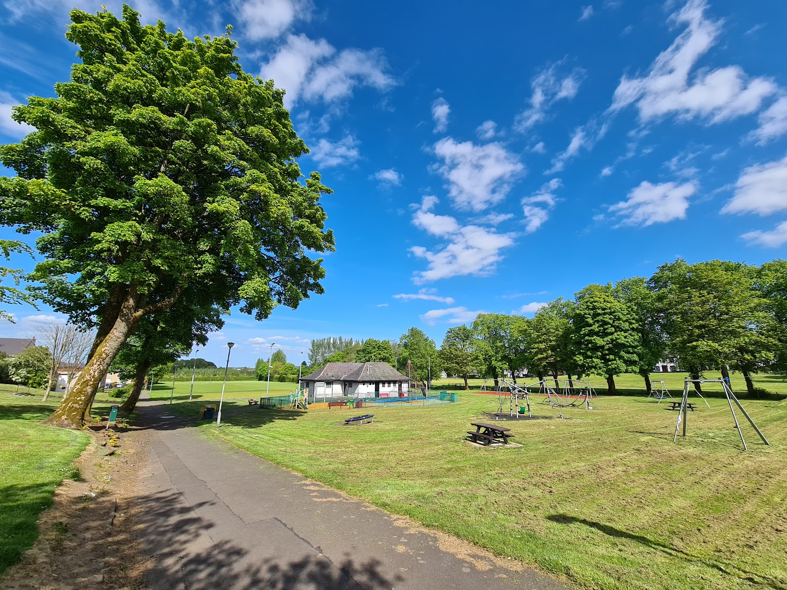 A path through a sunny village park in Eaglesham with picnic tables, a pavilion and tall trees.