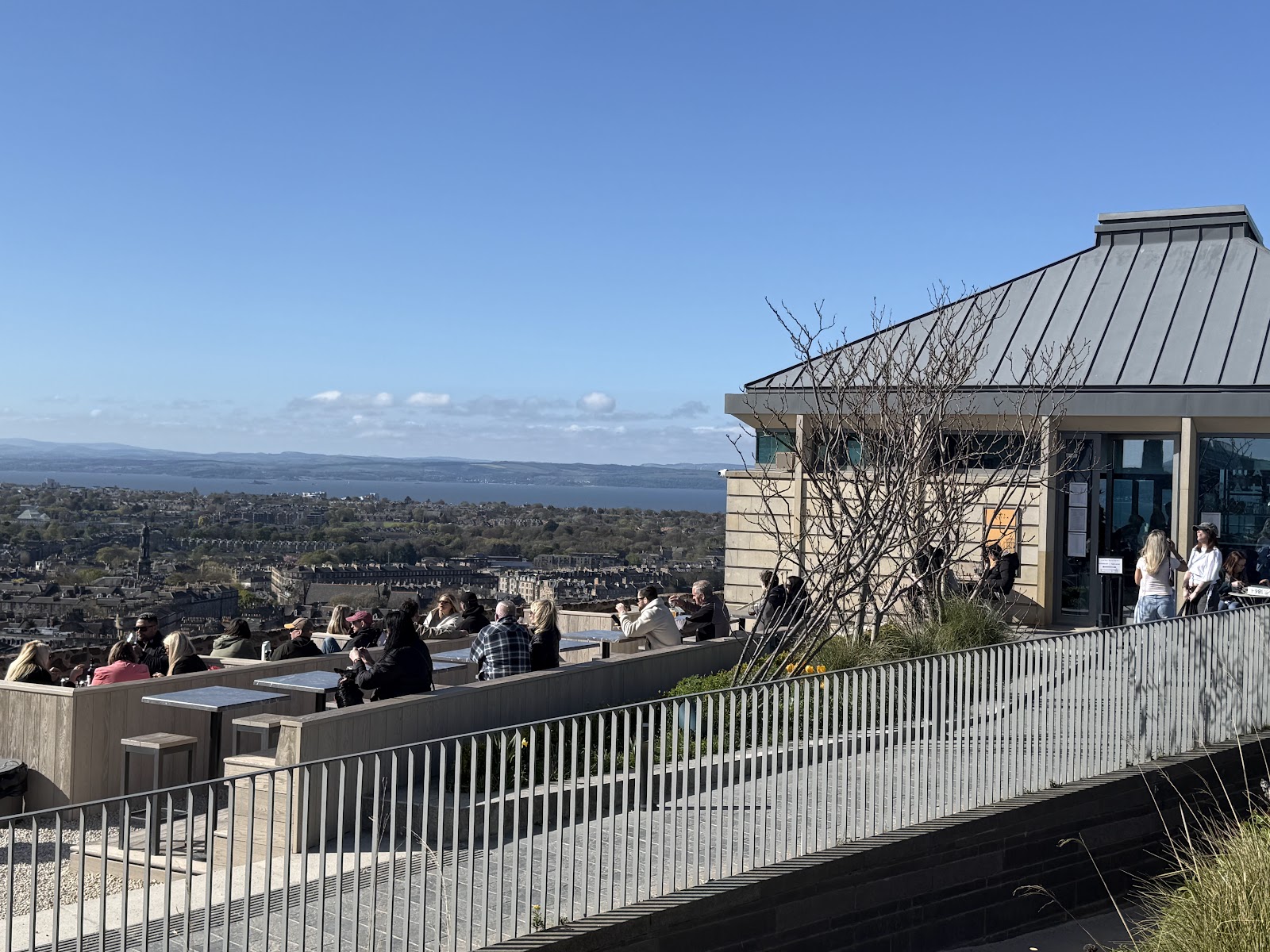 Cafe Calton's terrace on top of Calton Hill on a sunny day, people at tables looking out over the city to the Firth of Forth.
