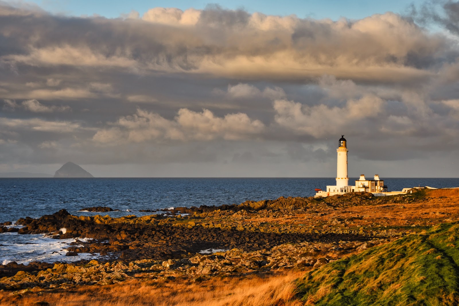 Corsewall Lighthouse's white tower and the old keepers' houses on the rocky point in low evening light, with Ailsa Craig on the horizon across the sea.