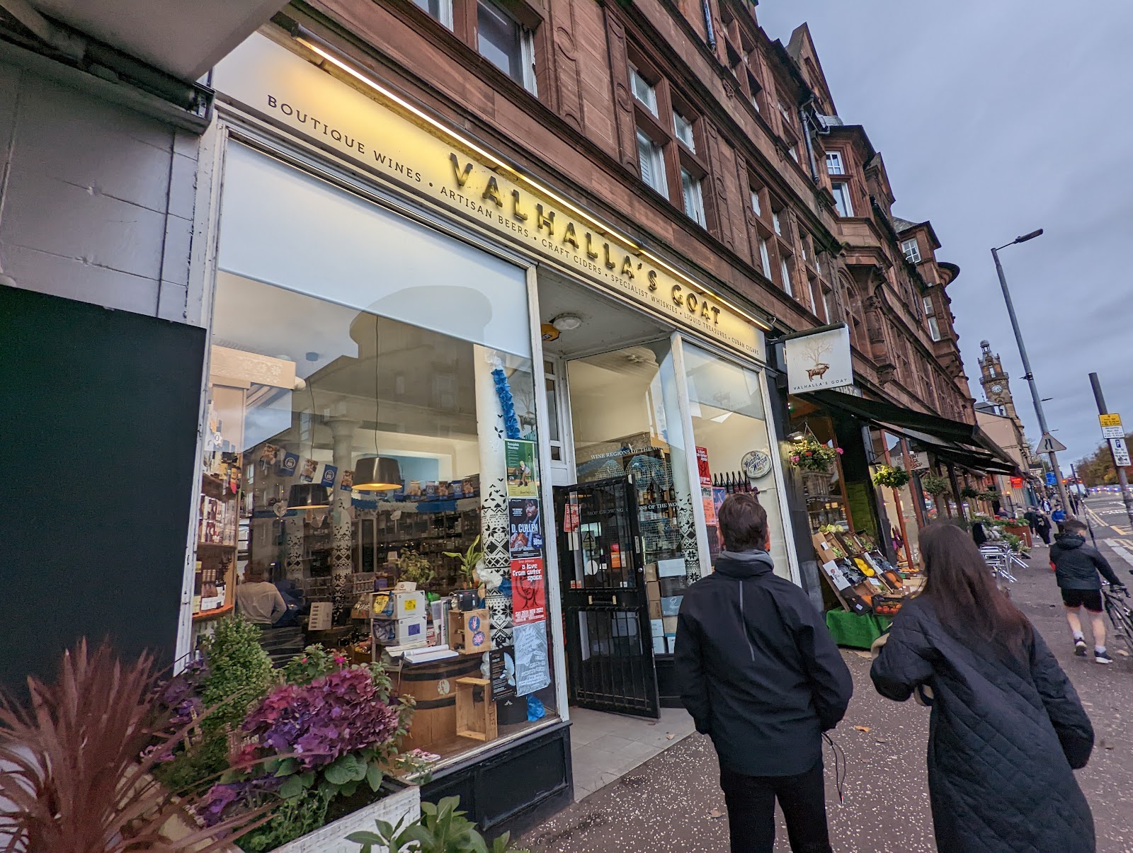 The front of Valhalla's Goat on Great Western Road, its long sign over windows of bottles, with plants by the door and people walking past.