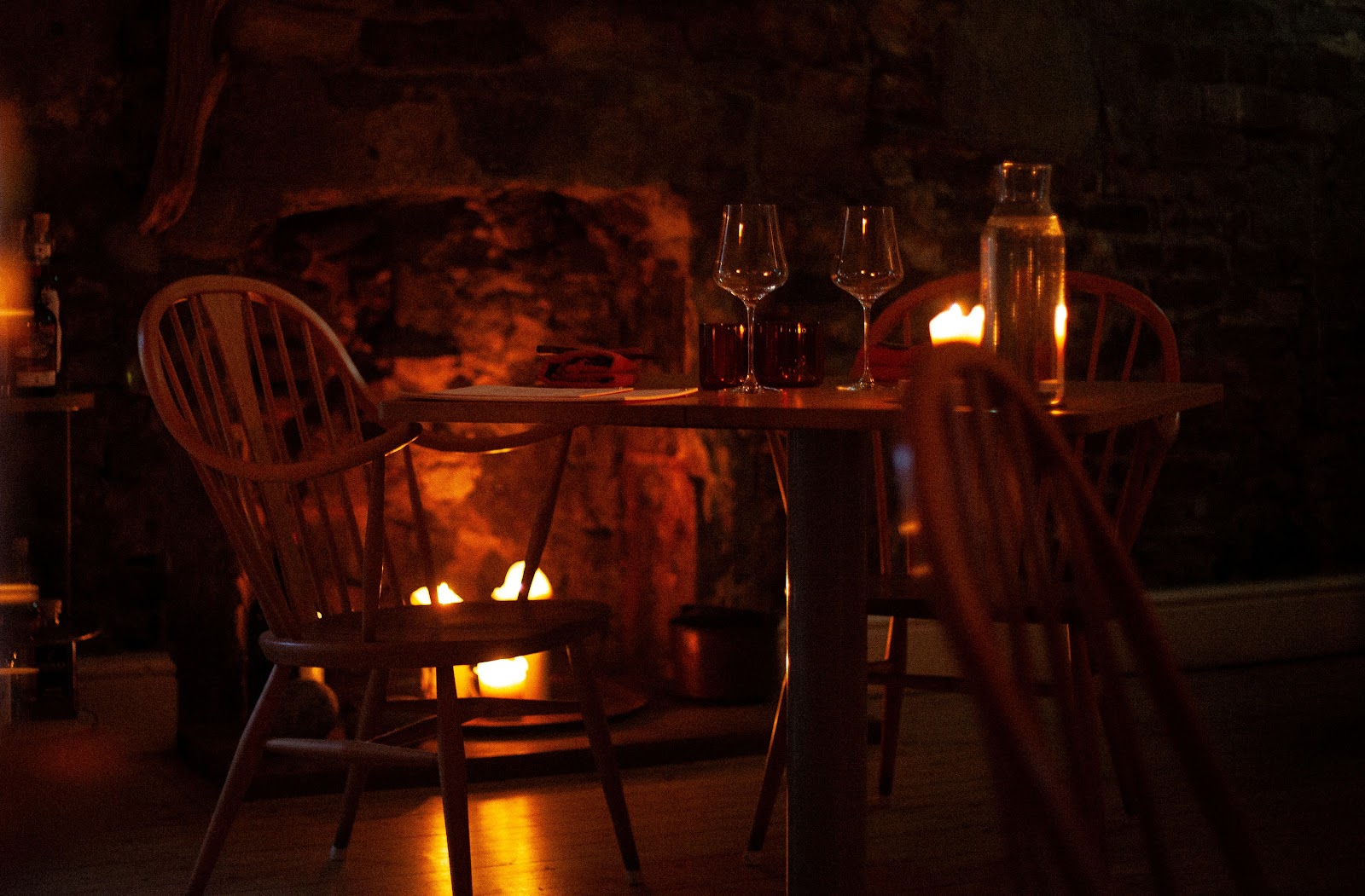 A table laid for two by candlelight in Montrose, with wooden chairs and a fire burning in the old stone fireplace behind.