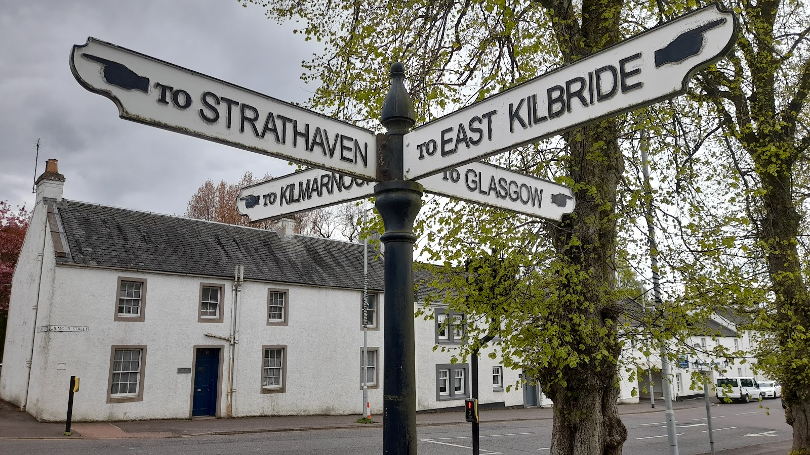 An old fingerpost in Eaglesham pointing to Strathaven, East Kilbride, Kilmarnock and Glasgow, cottages behind.