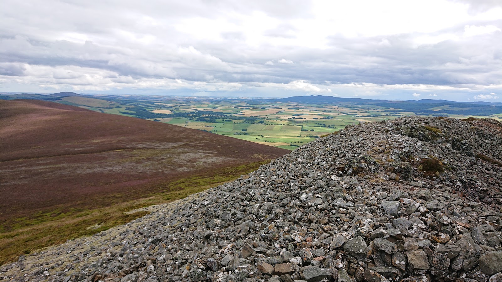 The bank of grey rubble that was the fort's wall on the summit of Tap o' Noth, with heather slopes and patchwork farmland running to distant hills below.