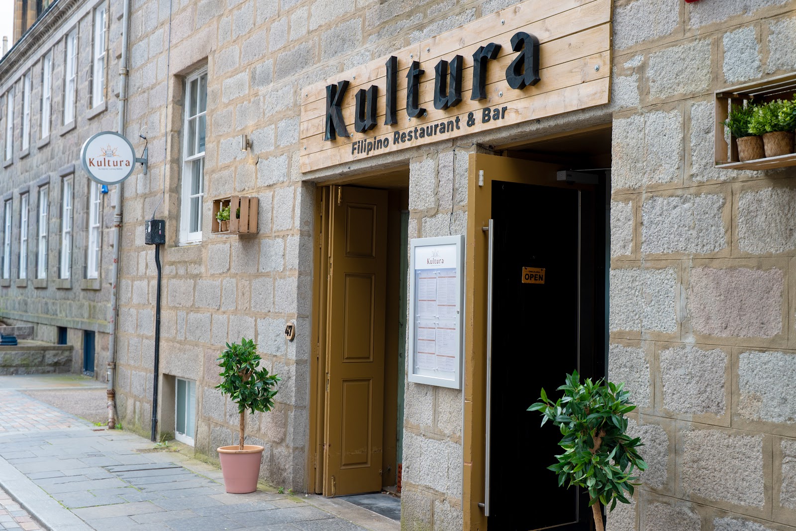 Kultura's front on Belmont Street, its wooden sign reading Filipino Restaurant & Bar above open doors in a granite wall, with potted bay trees by the step.