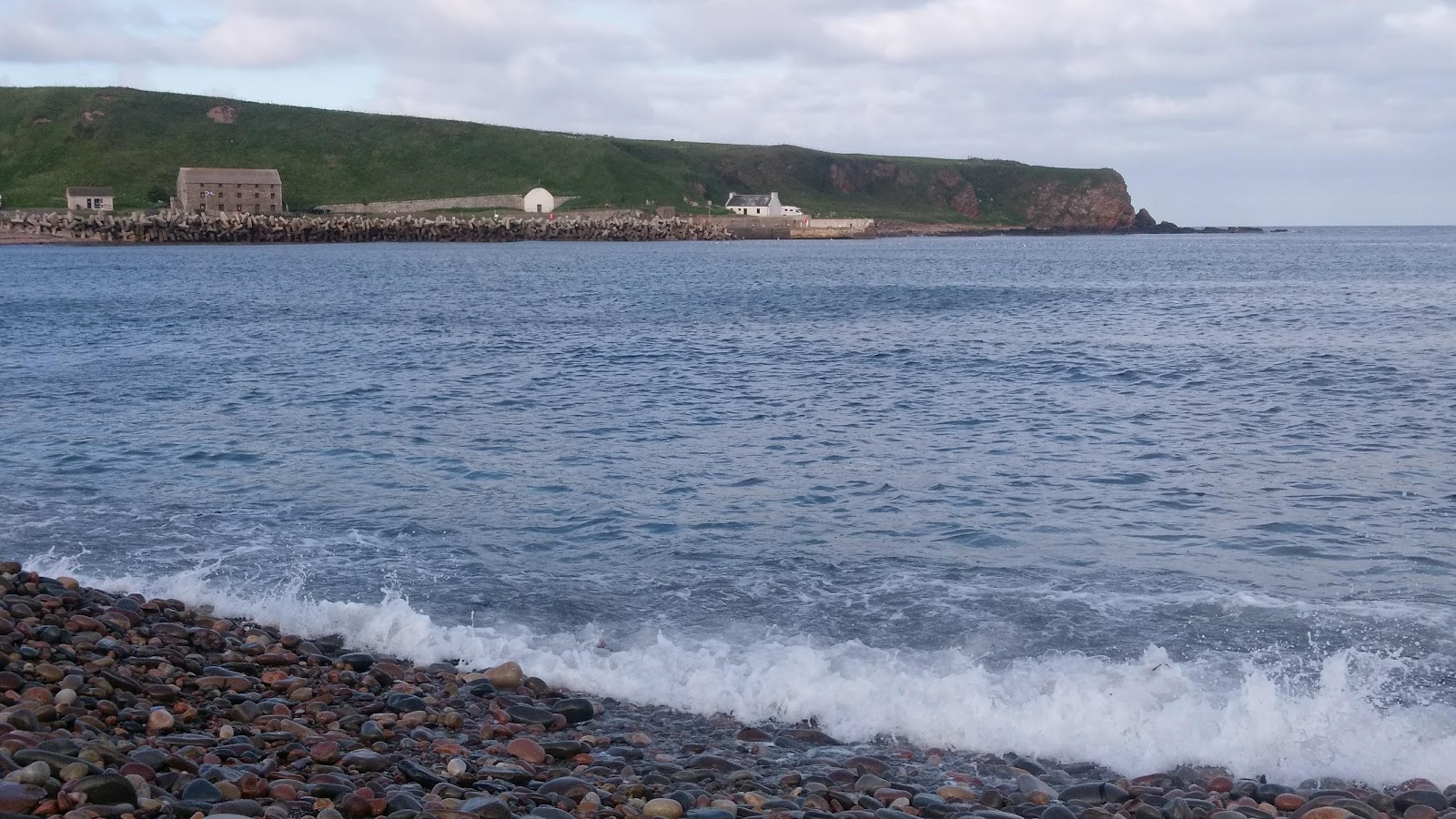 Dunbeath Bay, waves on a pebble shore and the harbour buildings below a green headland.