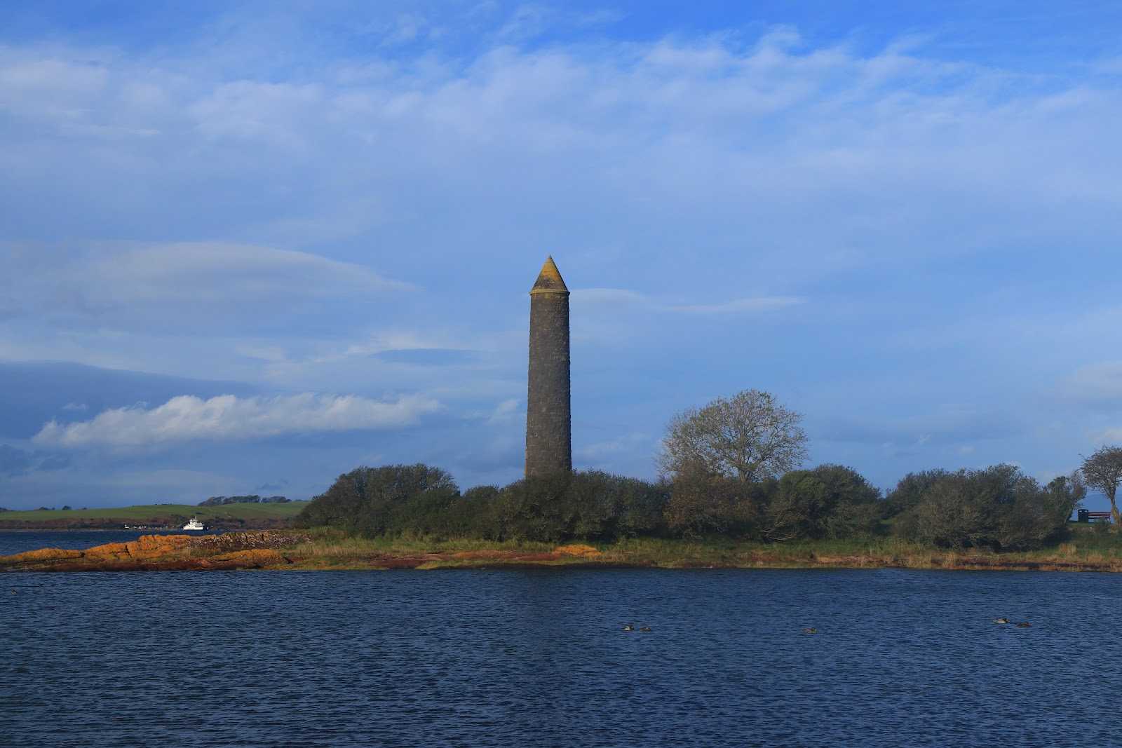 The Pencil at Largs, a tall round stone tower with a pointed top on the shore under a bright blue sky.