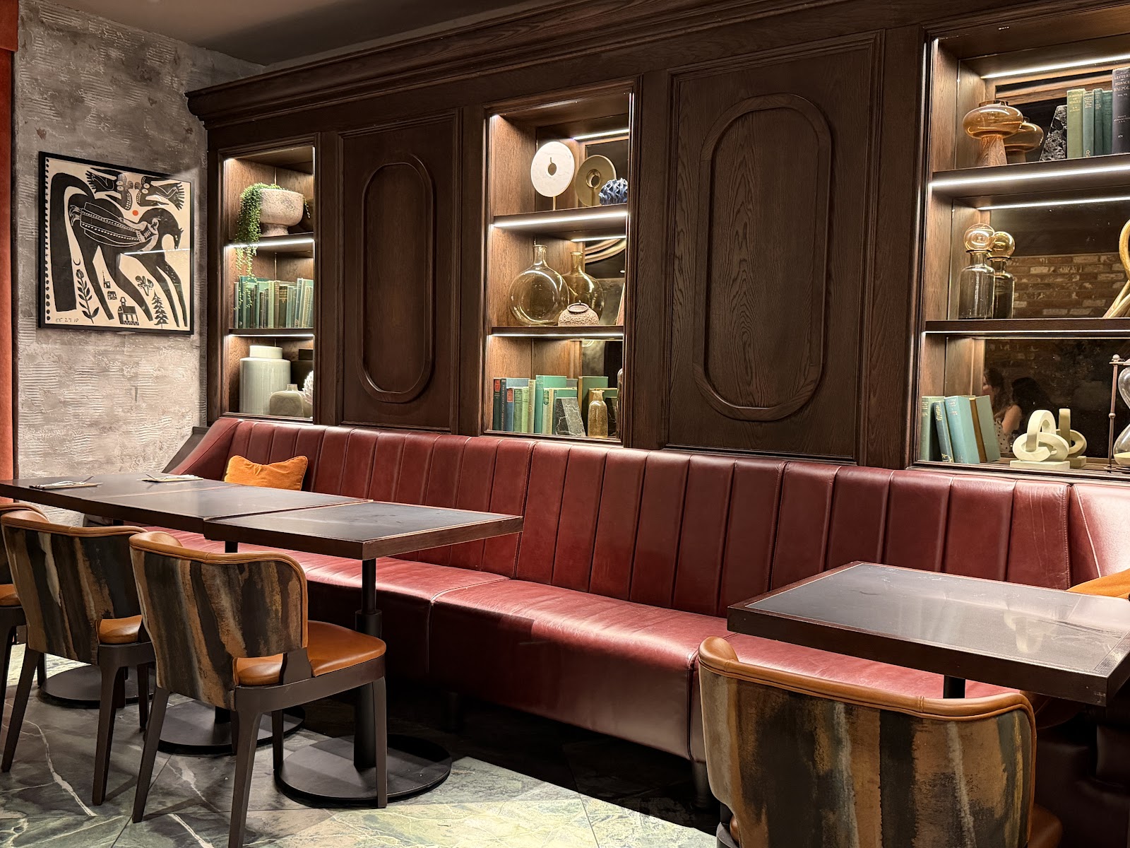 A long red leather banquette and dark tables in the dining room at Lennox, below lit shelves of books, vases and ornaments in dark wood panelling.