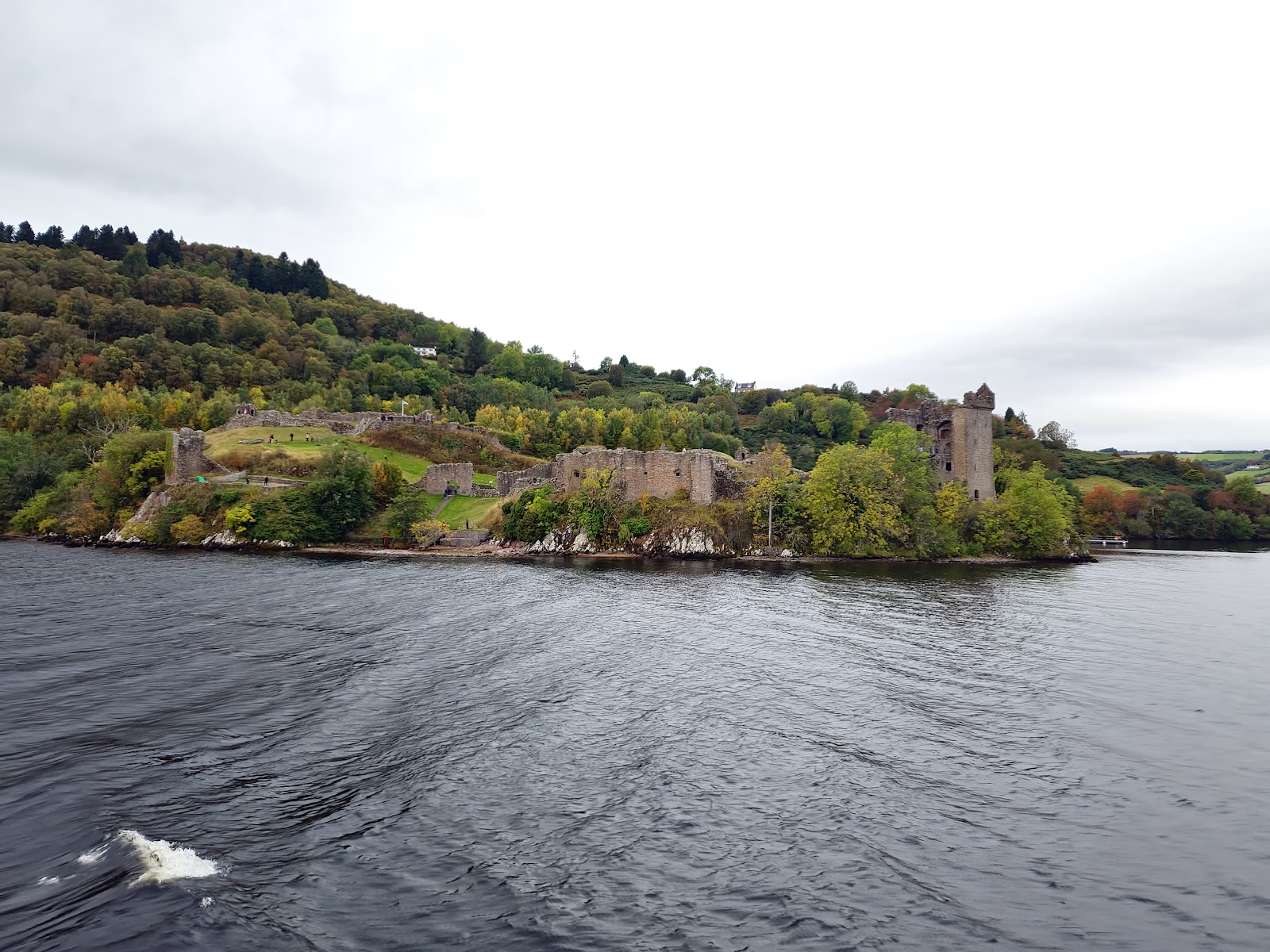 The ruins of Urquhart Castle on their wooded headland, seen across Loch Ness from the wake of a boat.