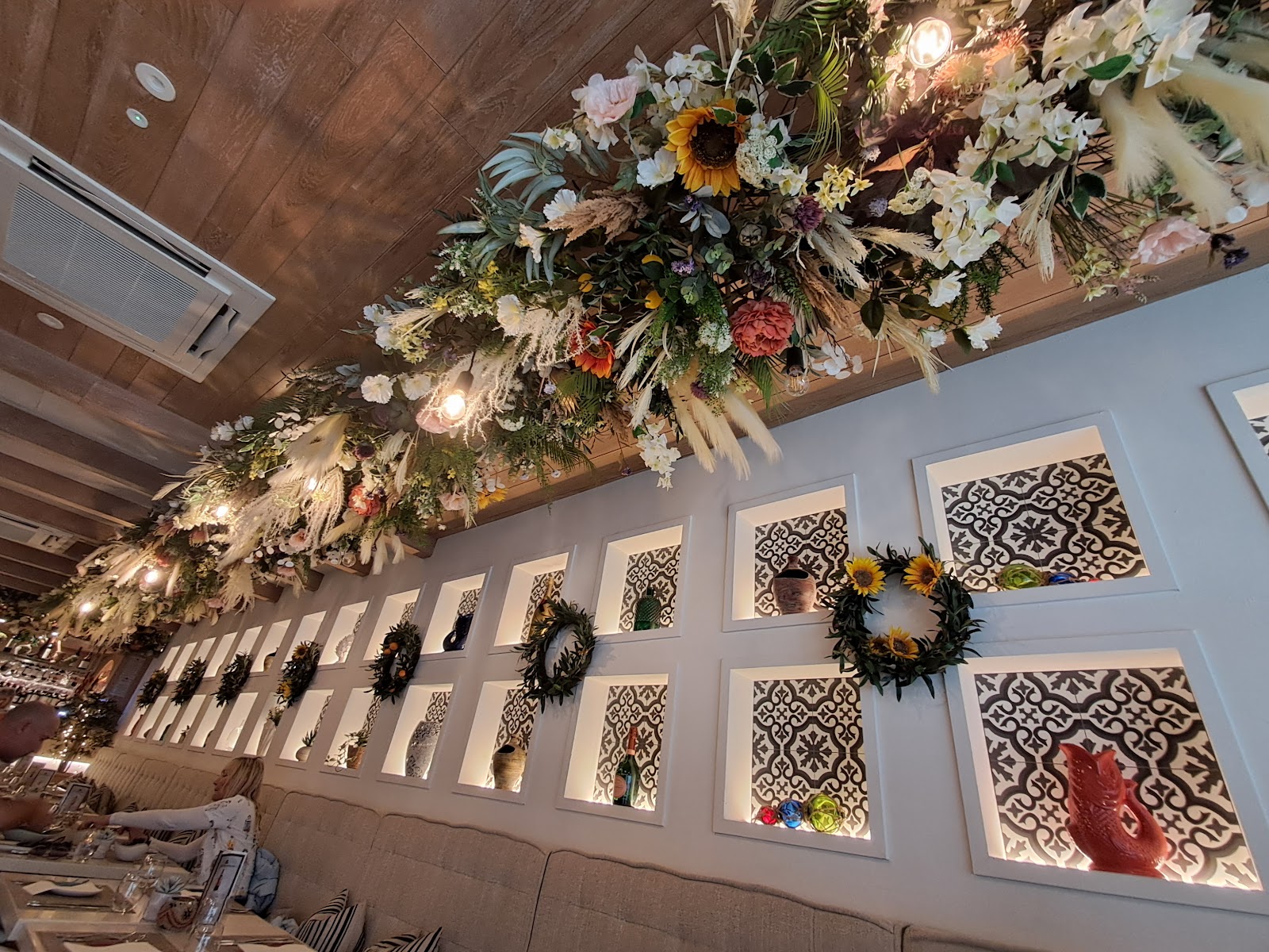 Inside Mediterranean on Thistle Street: a long ceiling garland of dried and silk flowers over a white wall of lit niches with patterned tiles, vases and wreaths.