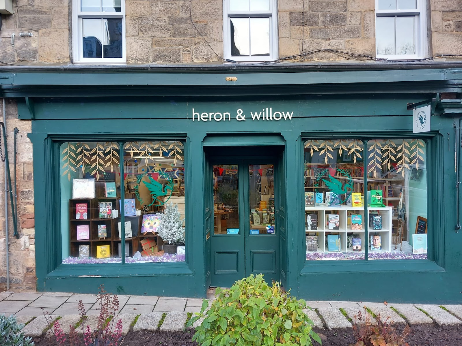 The deep green shopfront of Heron & Willow on the Canongate in Jedburgh, its two windows filled with books under painted willow leaves.
