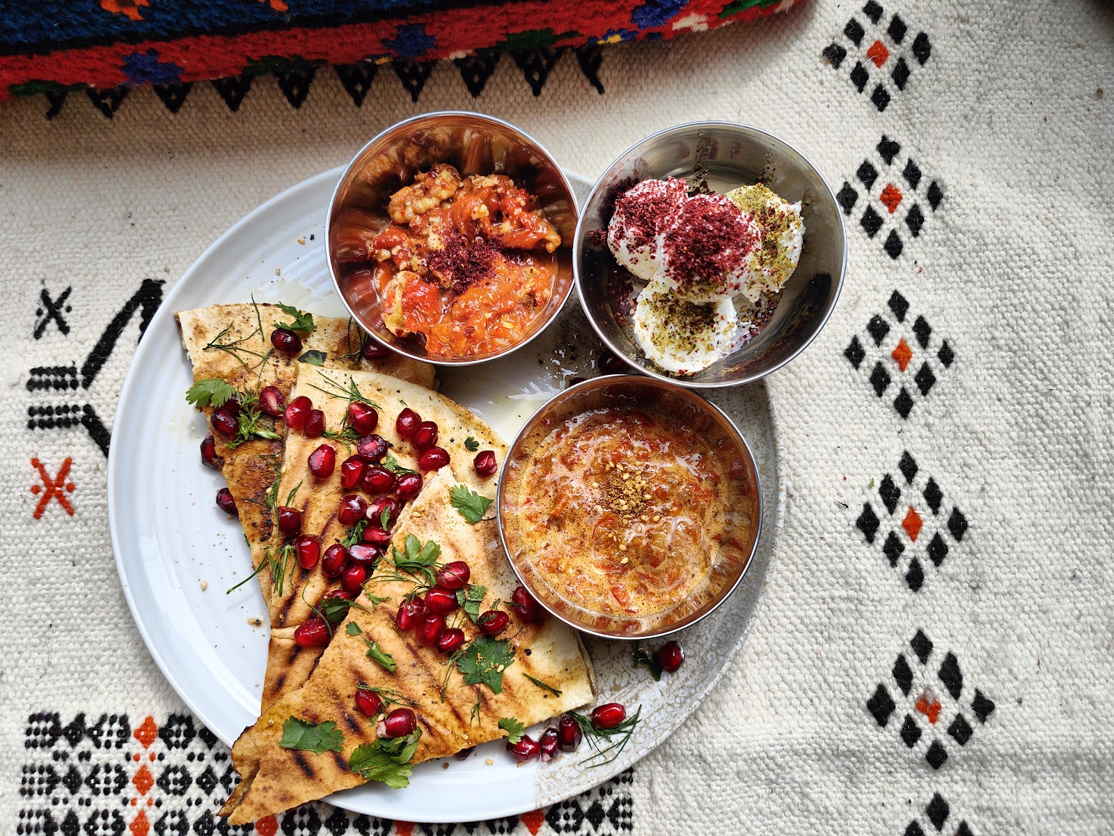 Grilled flatbread with pomegranate seeds and three dips in copper bowls, on a woven cloth at Qasba.