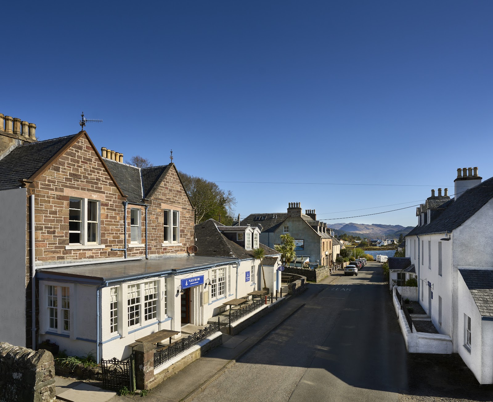 The Plockton Inn, a stone house with bay windows on a street in Plockton under a blue sky.