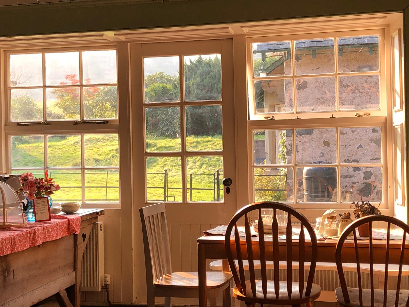 Low sun through the café's sash windows and glazed garden door, wooden chairs at the tables and green fields outside.