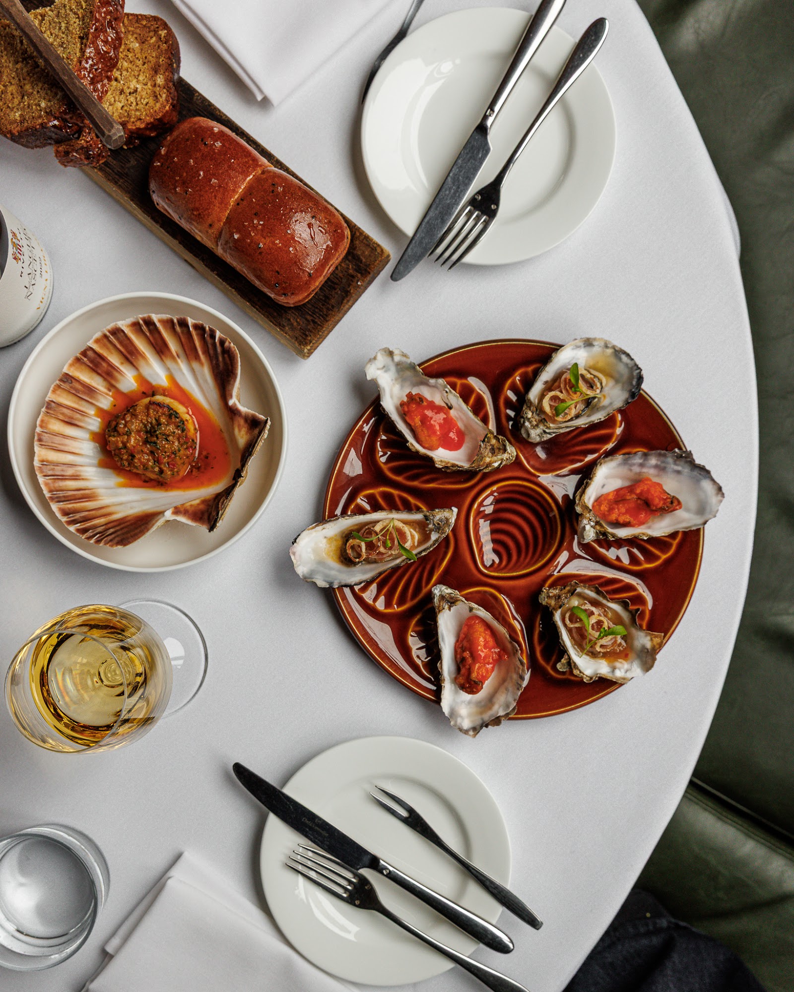 Oysters dressed two ways, a scallop shell, glazed milk bread and a glass of white wine on a table at Eleven Fifty Five.