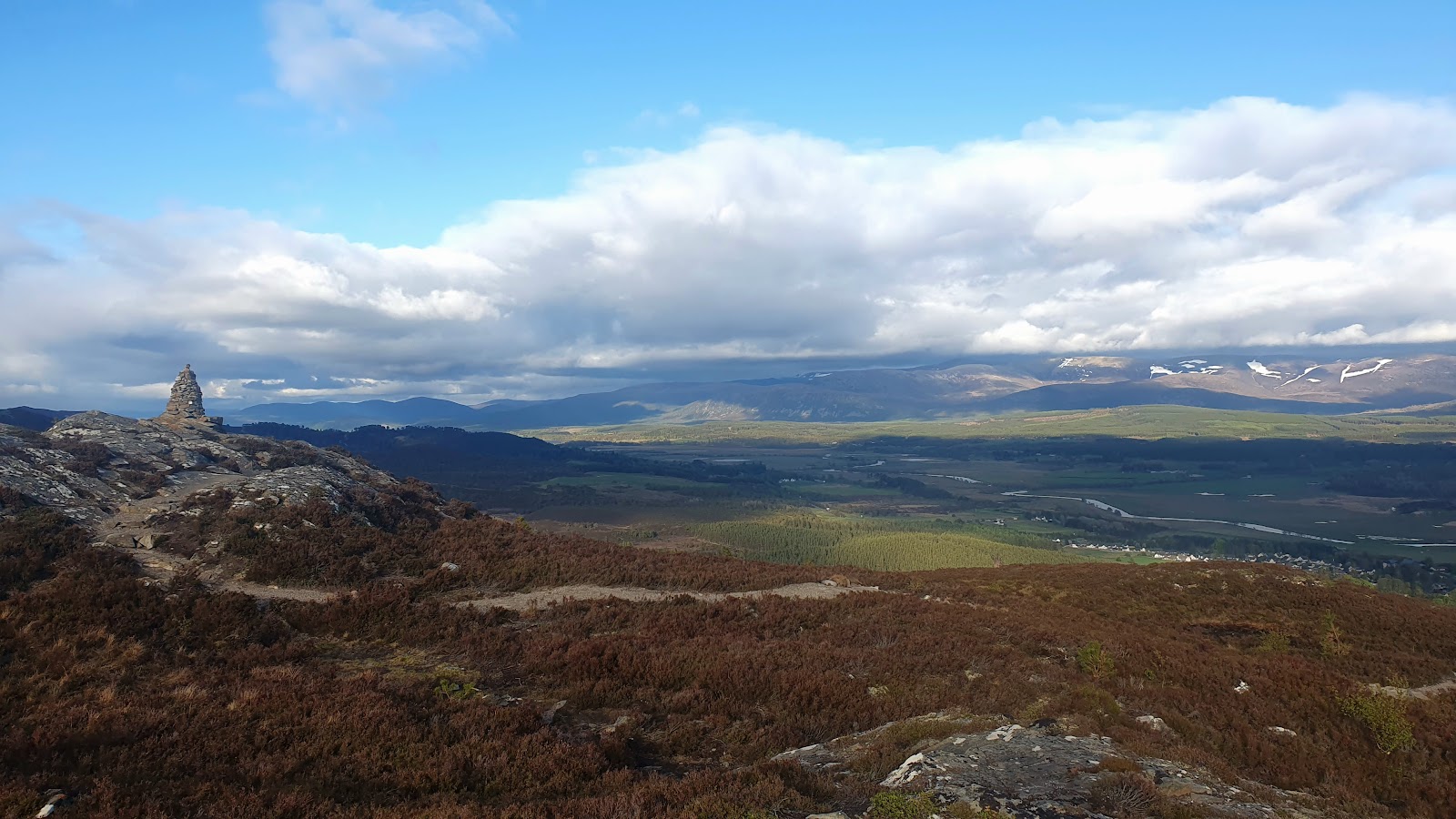 A tall cairn on the rocky crest of Creag Bheag, the path running through the heather and the Spey valley and the snow-streaked Cairngorms beyond.