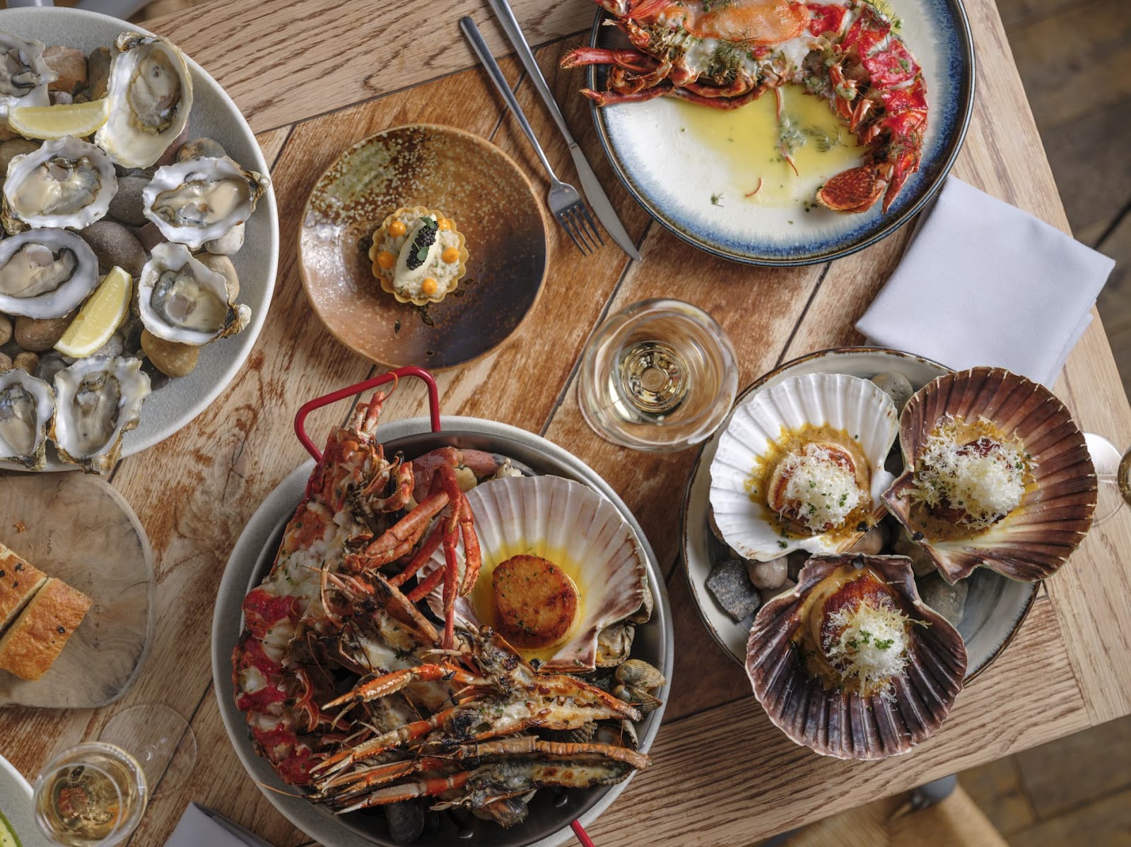 A wooden table at Dulse seen from above, laid with oysters on the half shell, grilled lobster, scallops in their shells and a pan of langoustines, with glasses of white wine.
