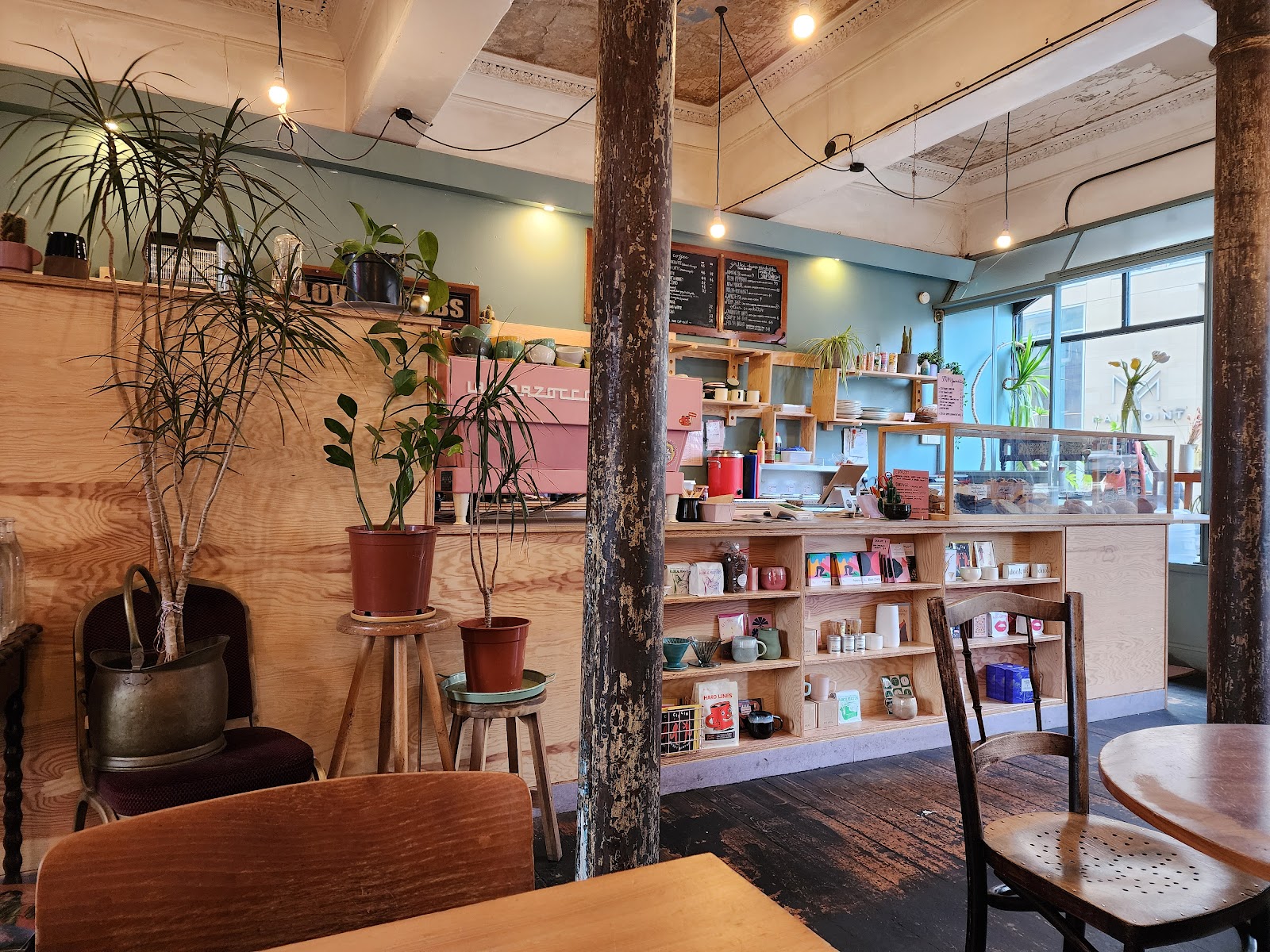 Inside Lovecrumbs on West Port: a plywood counter with a pink coffee machine, shelves of cups and coffee, plants, and old iron pillars.