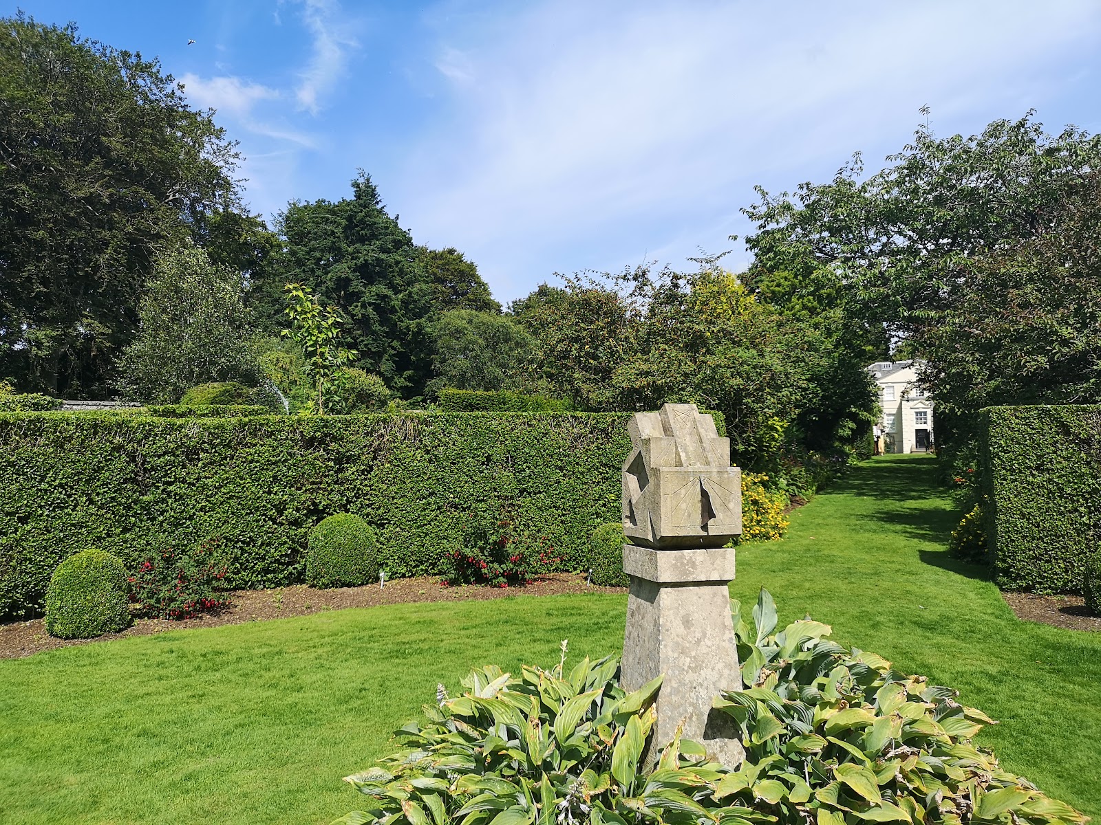 A stone sculpture on a lawn between tall yew hedges, with the white end of a building beyond.