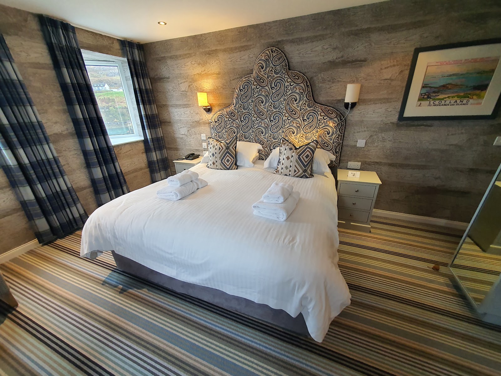 A bedroom at Hotel Hebrides: a white bed under a tall paisley headboard, tartan curtains and timber-effect walls, a window onto Tarbert.
