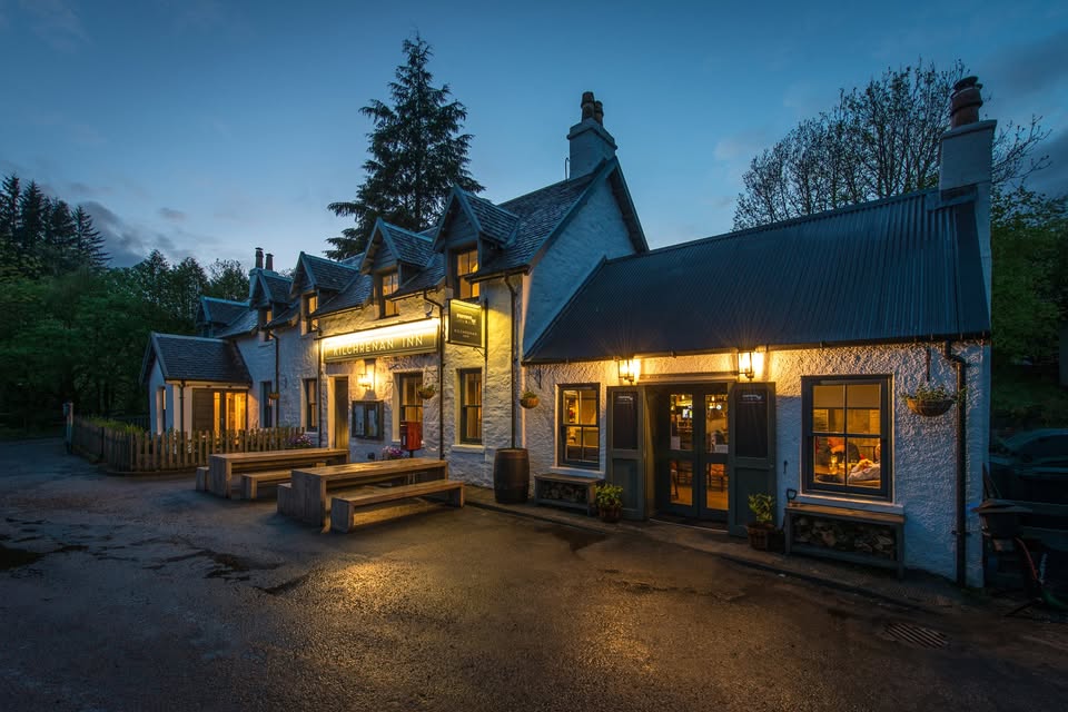 The Kilchrenan Inn at dusk, a long white building with its lamps lit, picnic benches out front and pine trees behind.