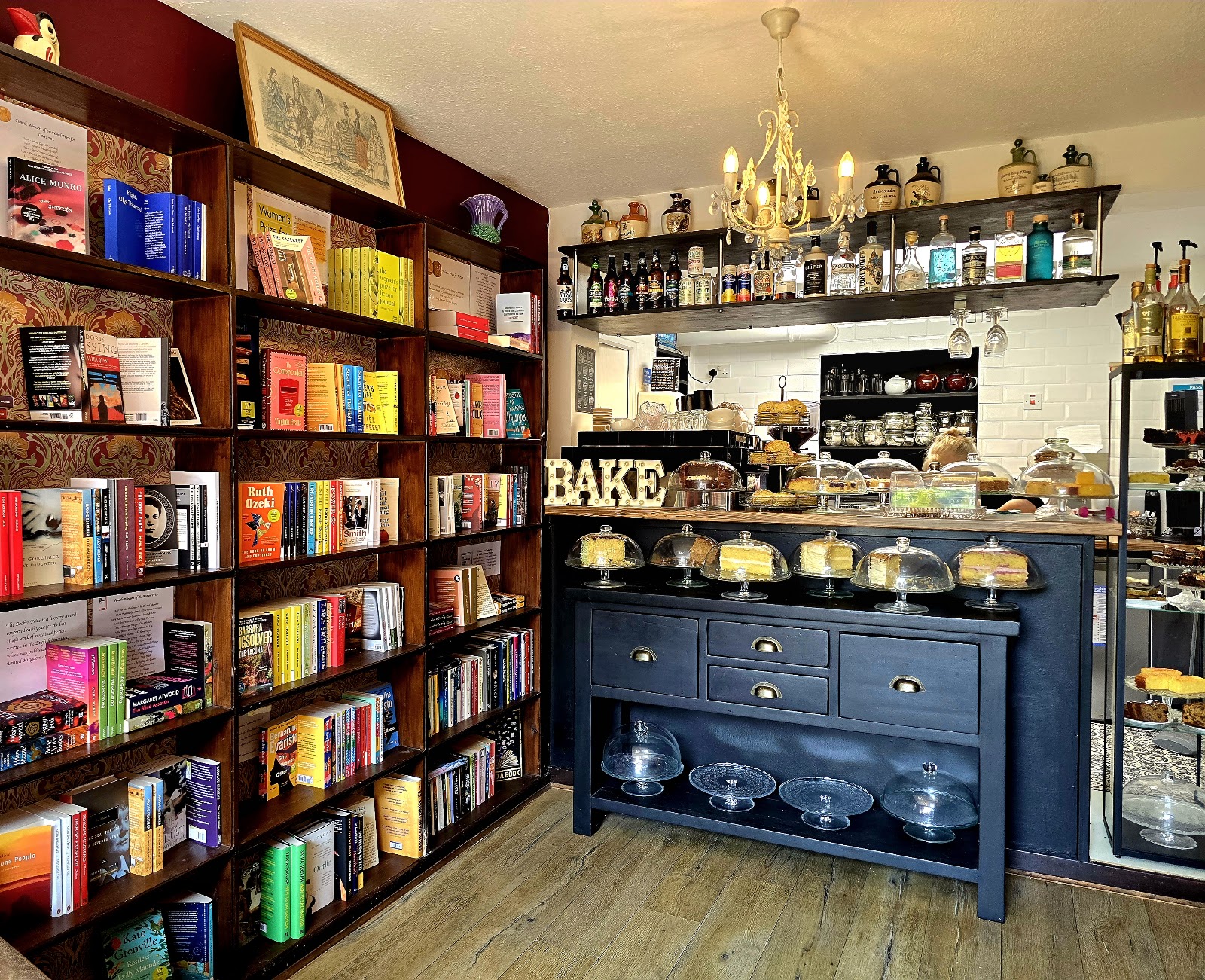 Inside ReadingLasses: shelves of new books by women writers on one side, and the café counter on the other, with cakes under glass domes, a chandelier and bottles on a shelf above.